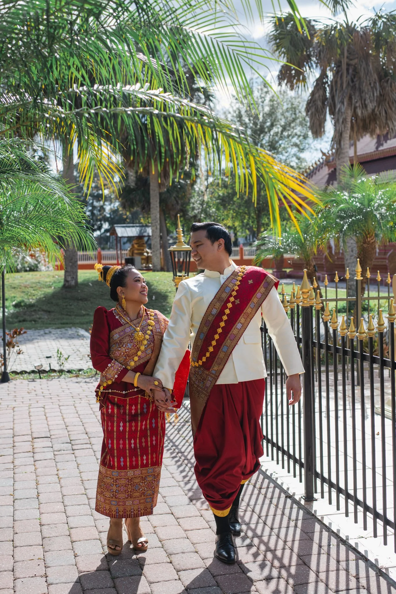 A Southeast Asian couple in traditional wedding attire walking and holding hands outdoors with palm trees and a temple in the background.