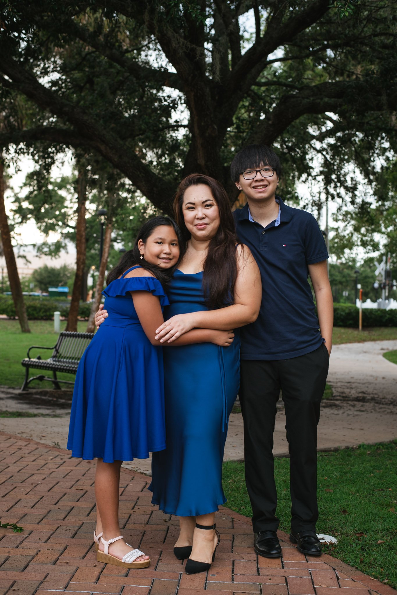 A woman and two children standing outdoors under a large tree. The woman is wearing a blue dress, the girl is wearing a matching blue dress, and the boy is wearing a dark blue shirt and black pants.