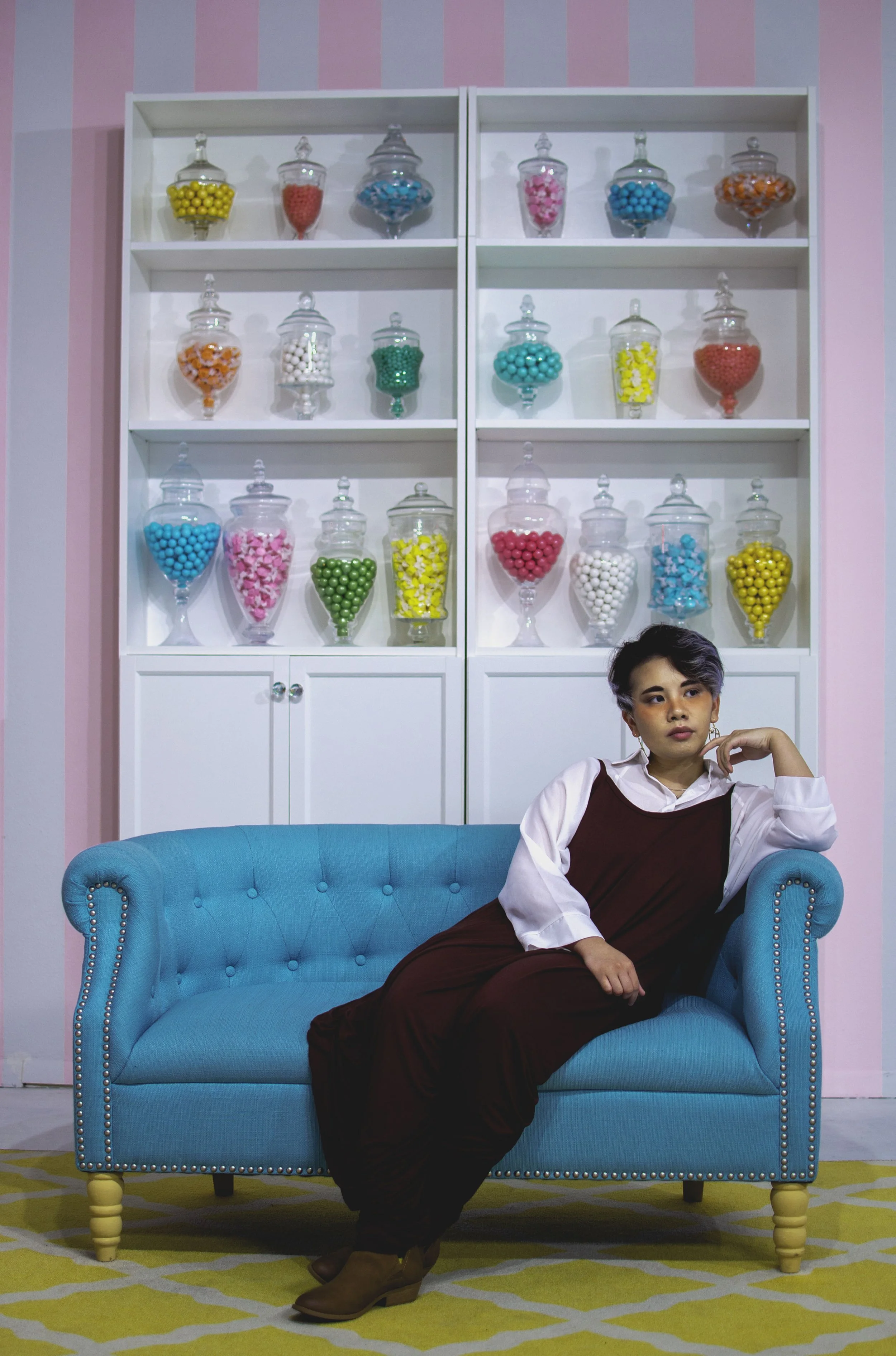 A woman with short hair, wearing a white blouse and maroon dress, sitting on a bright blue tufted sofa, with a colorful candy display behind her, in a room with pink and blue striped walls and a yellow patterned rug.