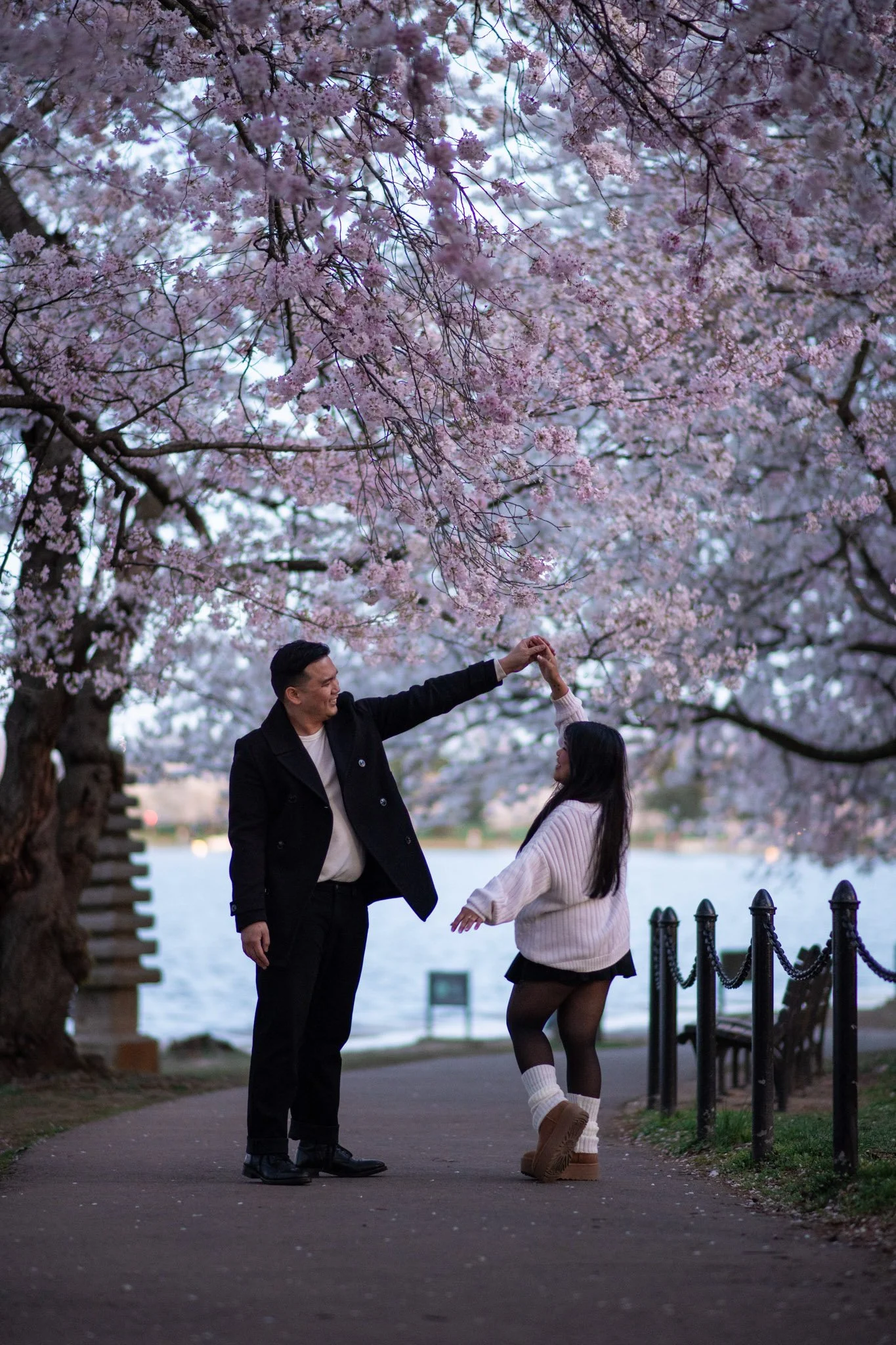 A man and woman dancing together under pink cherry blossom trees near a body of water, with benches and chains along the walkway.