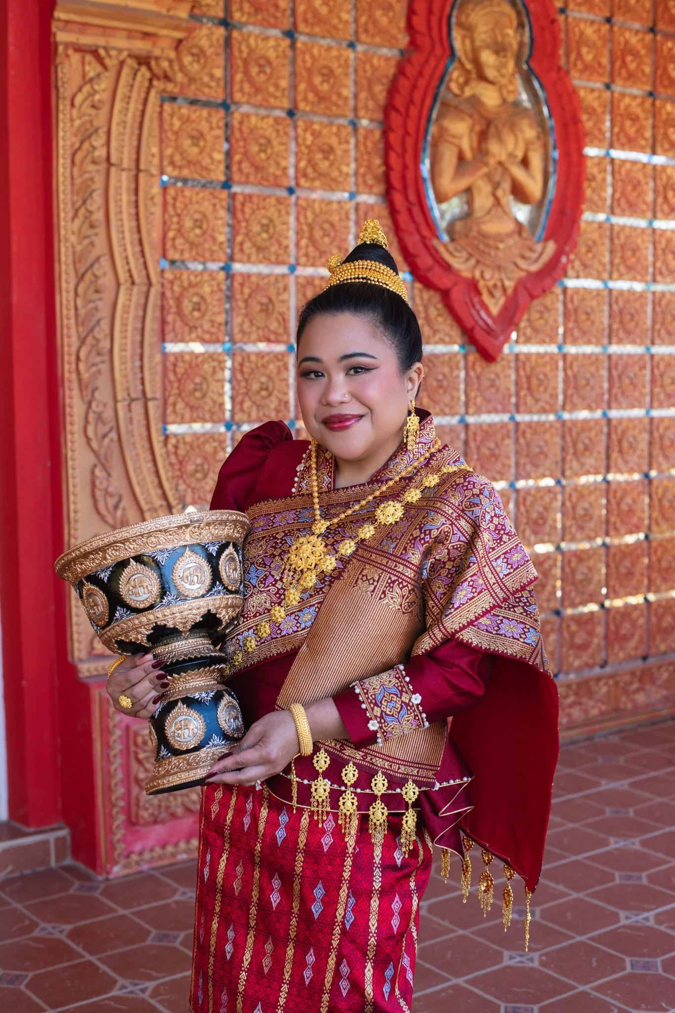 A woman in traditional Southeast Asian attire, holding a decorated ceremonial bowl, standing in front of an ornate wall with a carved image of a deity or figure, jewelry, and rich fabric details.