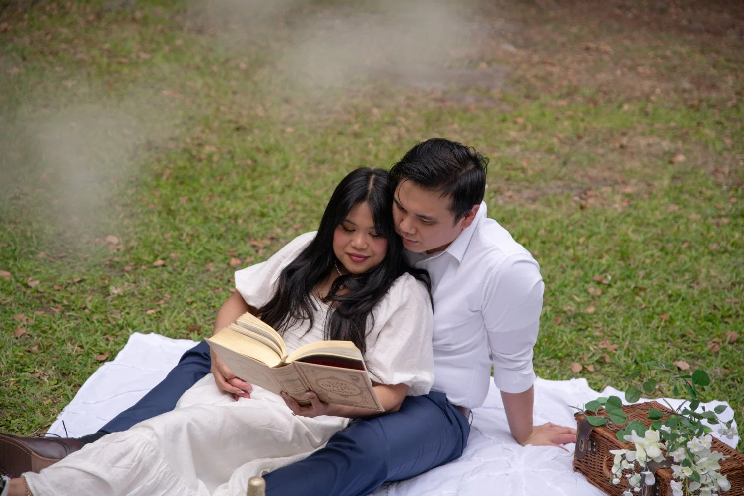 A couple sitting on a white blanket outdoors on grass, reading a book together, with a basket of flowers nearby.