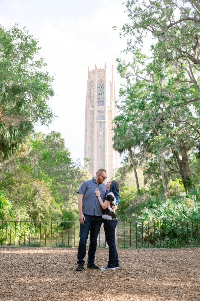 A couple standing close together in a wooded park with a tall, historic tower in the background, near a body of water surrounded by trees.