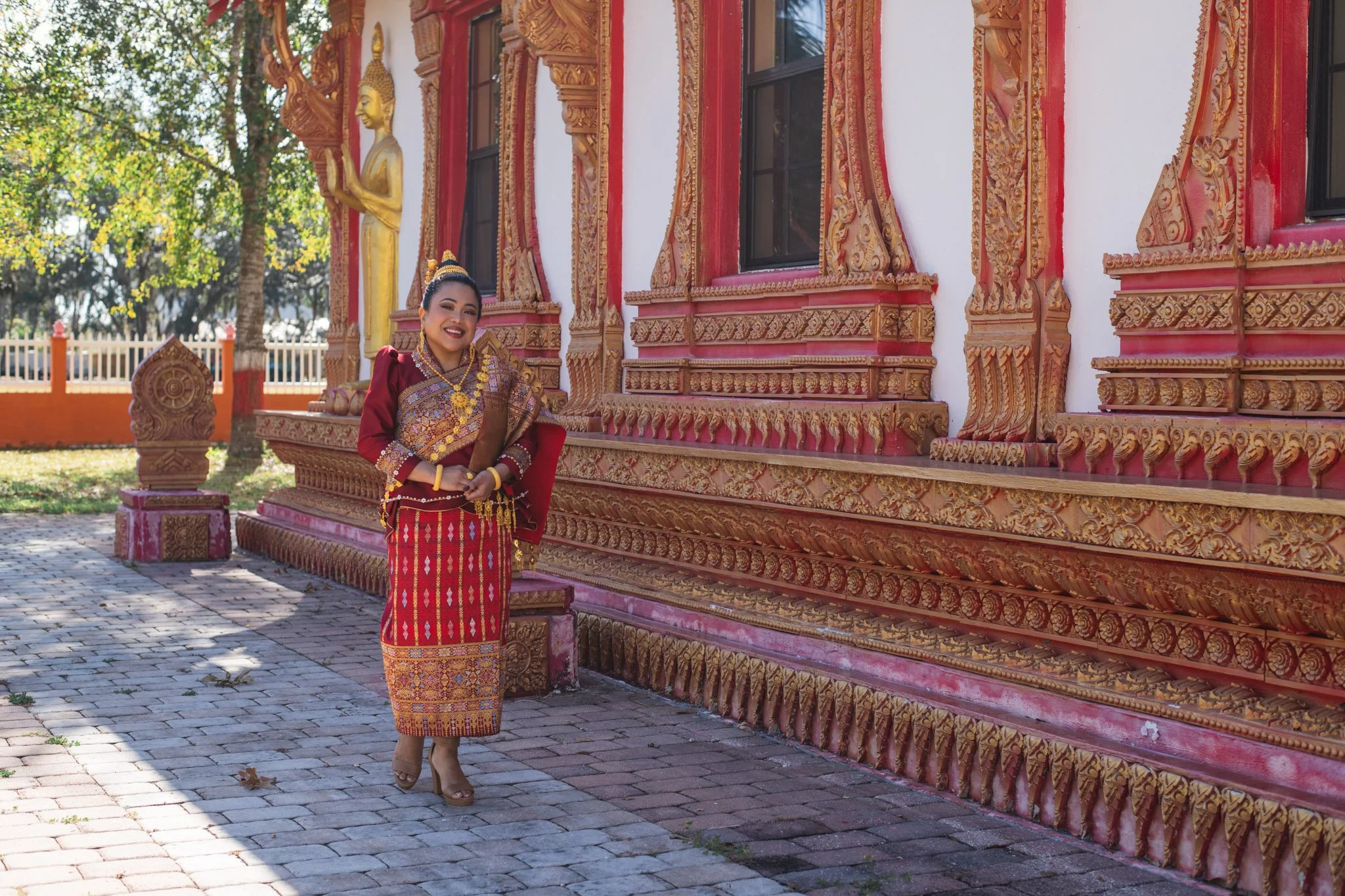 A woman dressed in traditional Southeast Asian attire, standing outside a decorated temple with ornate wood carvings and gold accents.