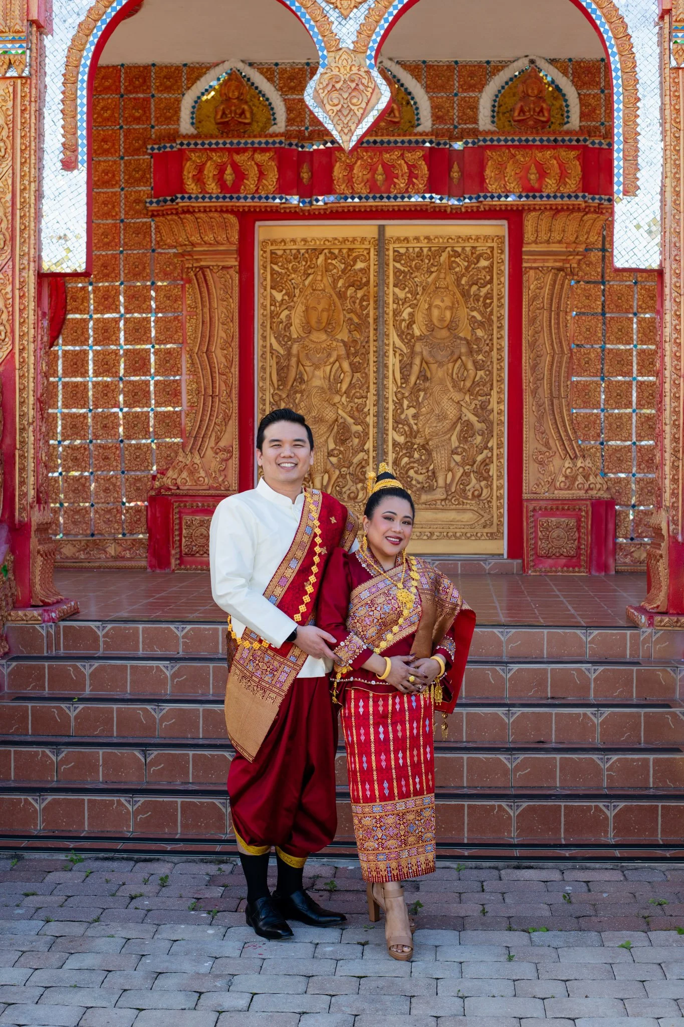 A couple dressed in traditional Thai attire standing in front of an ornately decorated temple door with golden carvings.