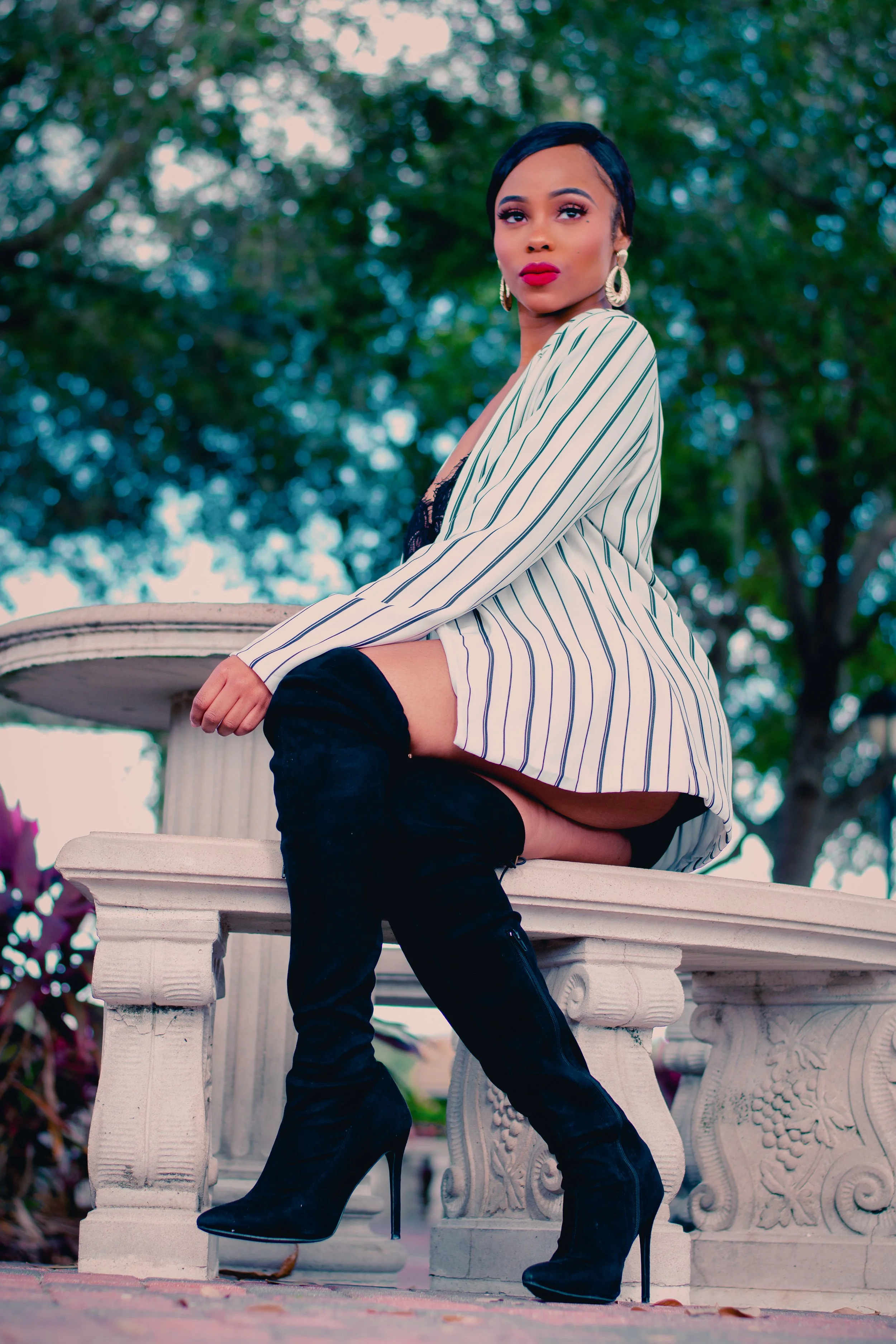 A woman in a striped blazer, black lace top, and thigh-high black boots sitting on a stone bench outdoors, with trees in the background.