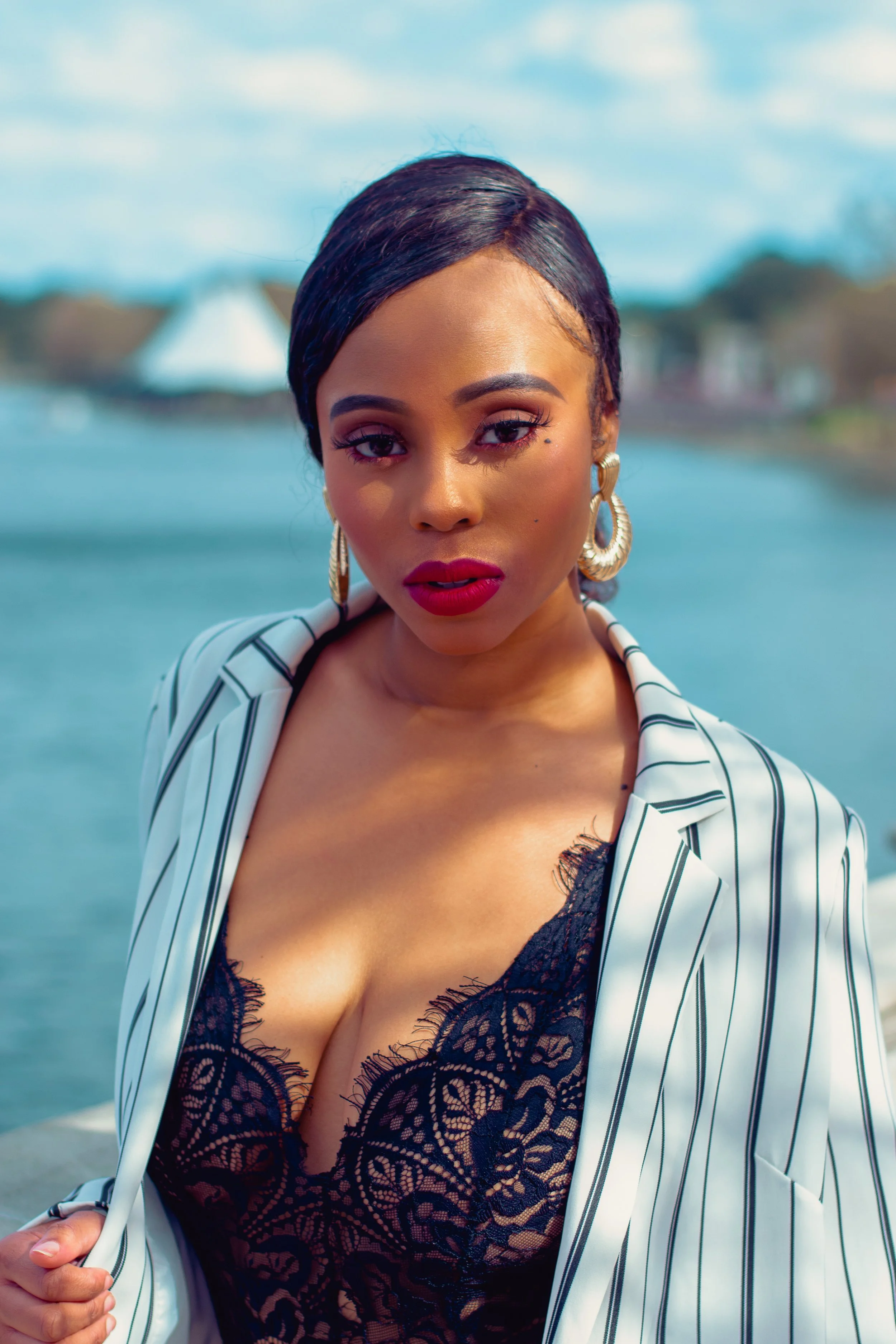 A woman with short black hair, wearing large gold hoop earrings, a lace black top, a white blazer with black pinstripes, and red lipstick, standing outdoors near a body of water with houses in the background.