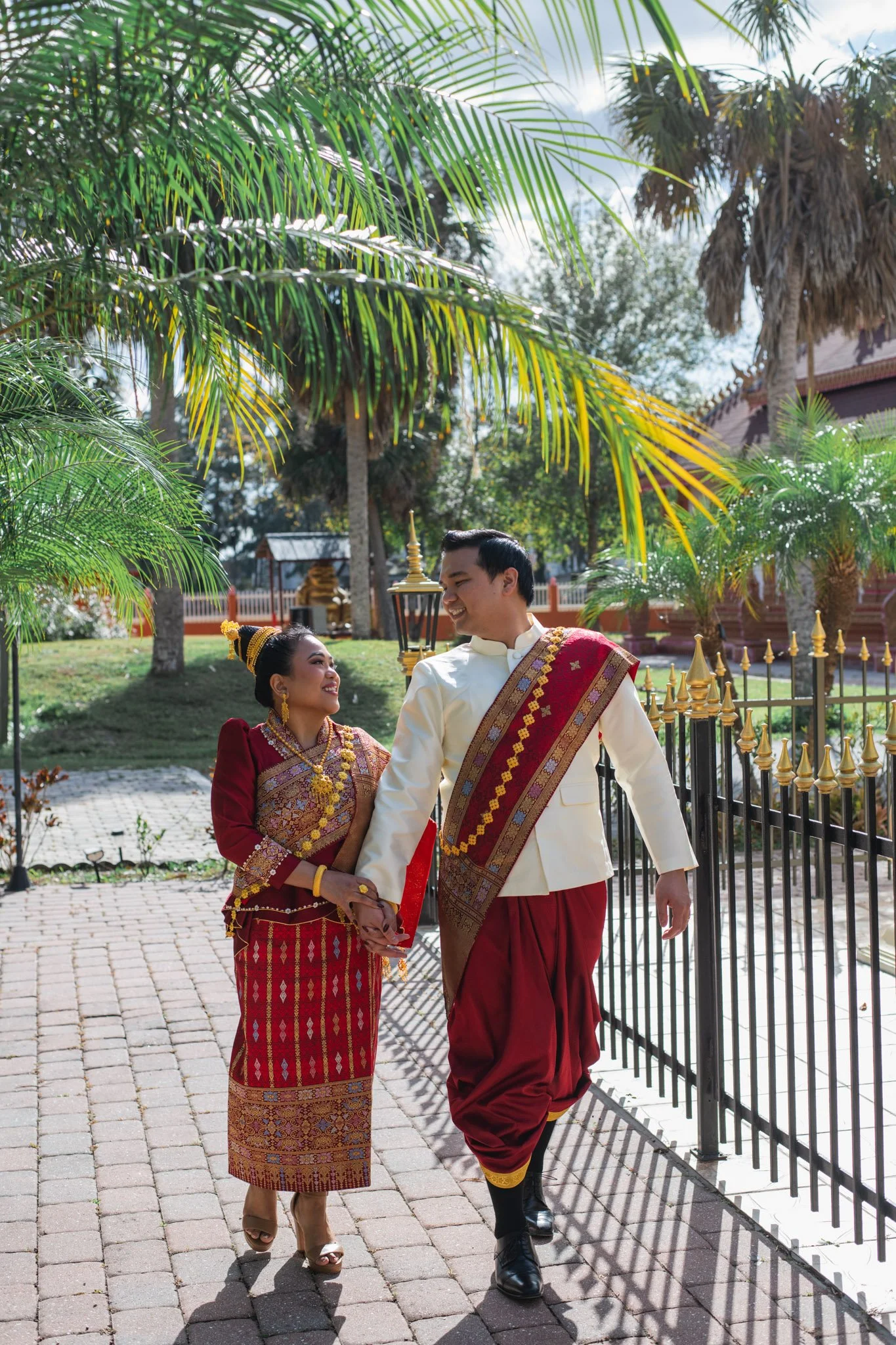 A couple dressed in traditional Southeast Asian attire walking hand in hand outdoors, surrounded by lush green trees and a sunny sky.