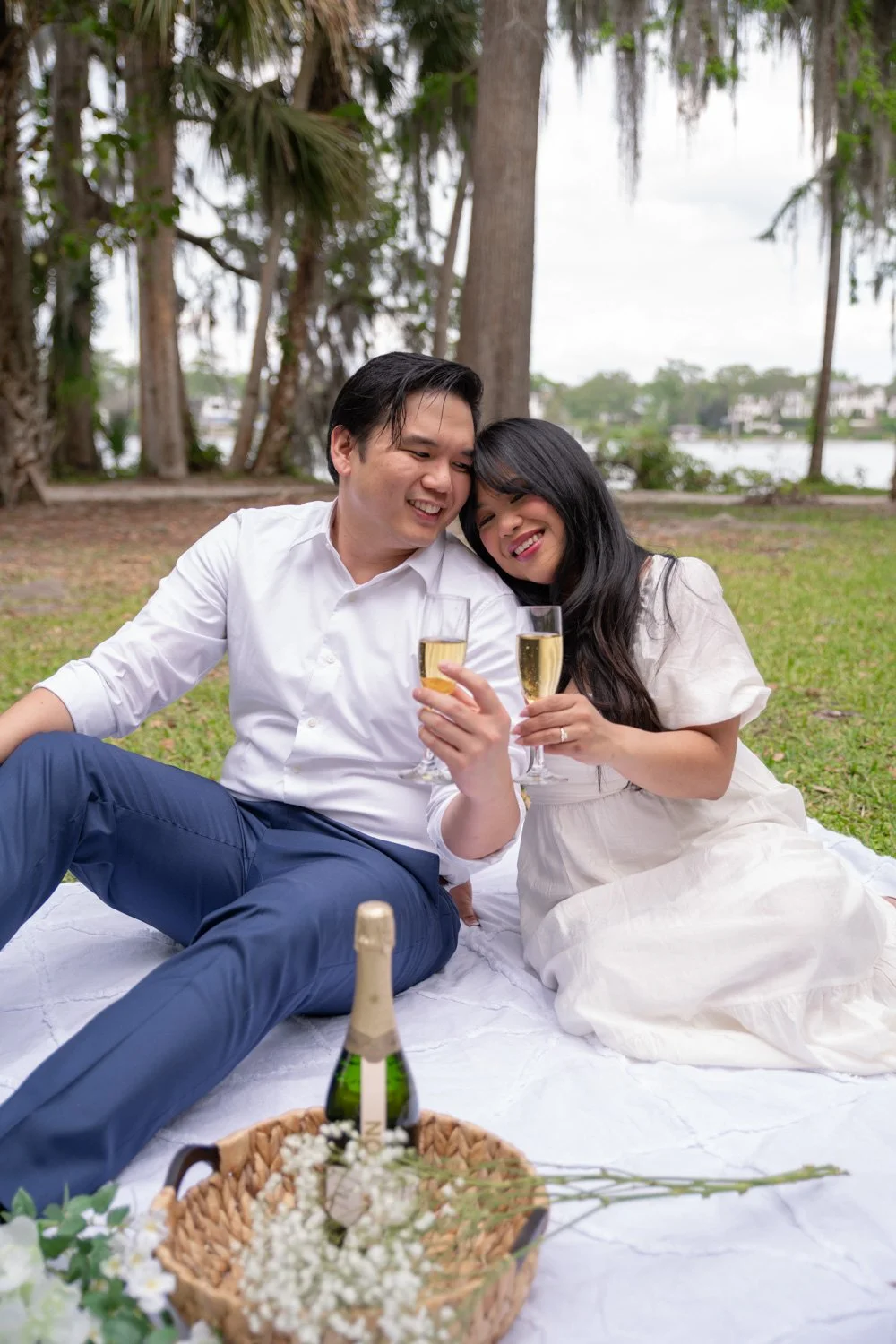 A couple having a romantic picnic outdoors, sitting on a white blanket, holding champagne glasses, smiling and leaning on each other, with a basket of flowers and a champagne bottle in front of them, surrounded by trees near a river.