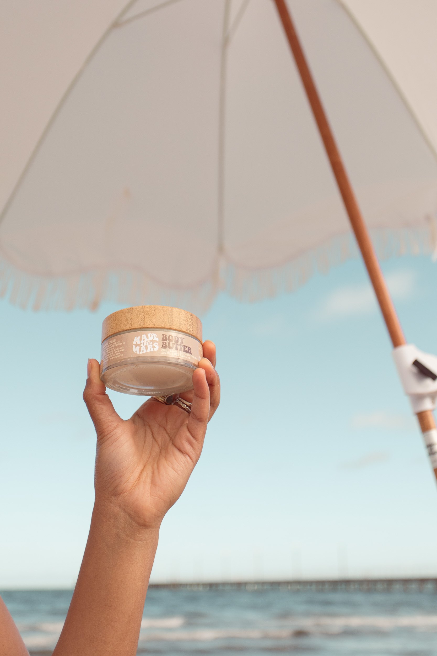 hand holding moisturiser in air against umbrella at beach photoshoot