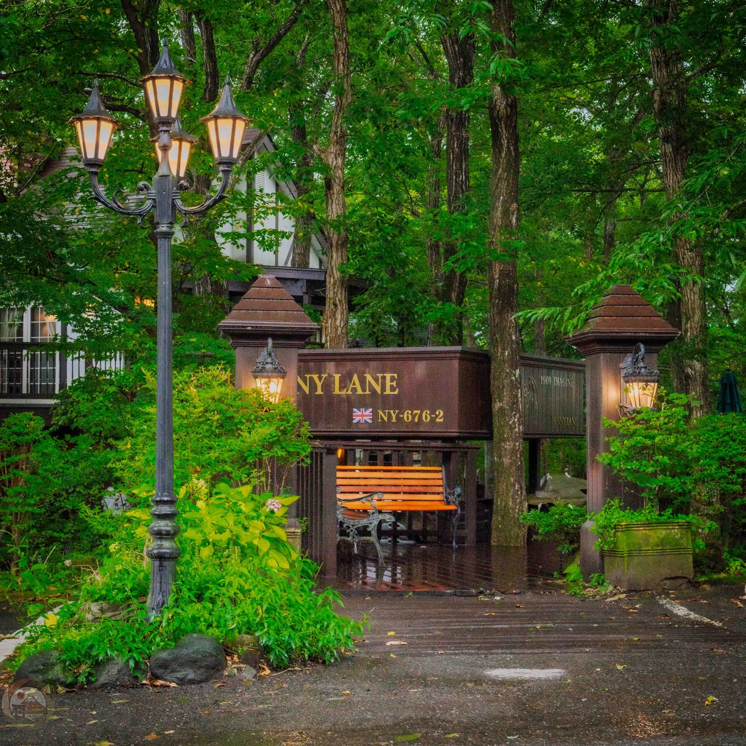 Entrance to Penny Lane Bakery surrounded by dense green trees, with warm street lamps glowing beside a wooden bench and wet ground after rainfall.