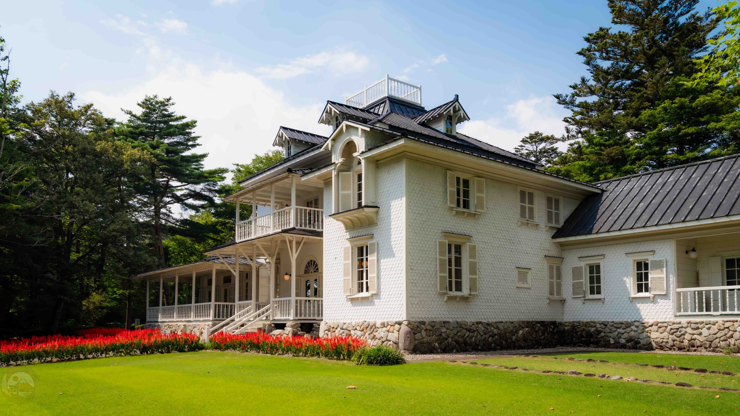 A historic white German-inspired villa in Tochigi, Japan, featuring wooden balconies, shuttered windows, and a stone foundation, surrounded by bright red flowers and lush green trees under a sunny blue sky.