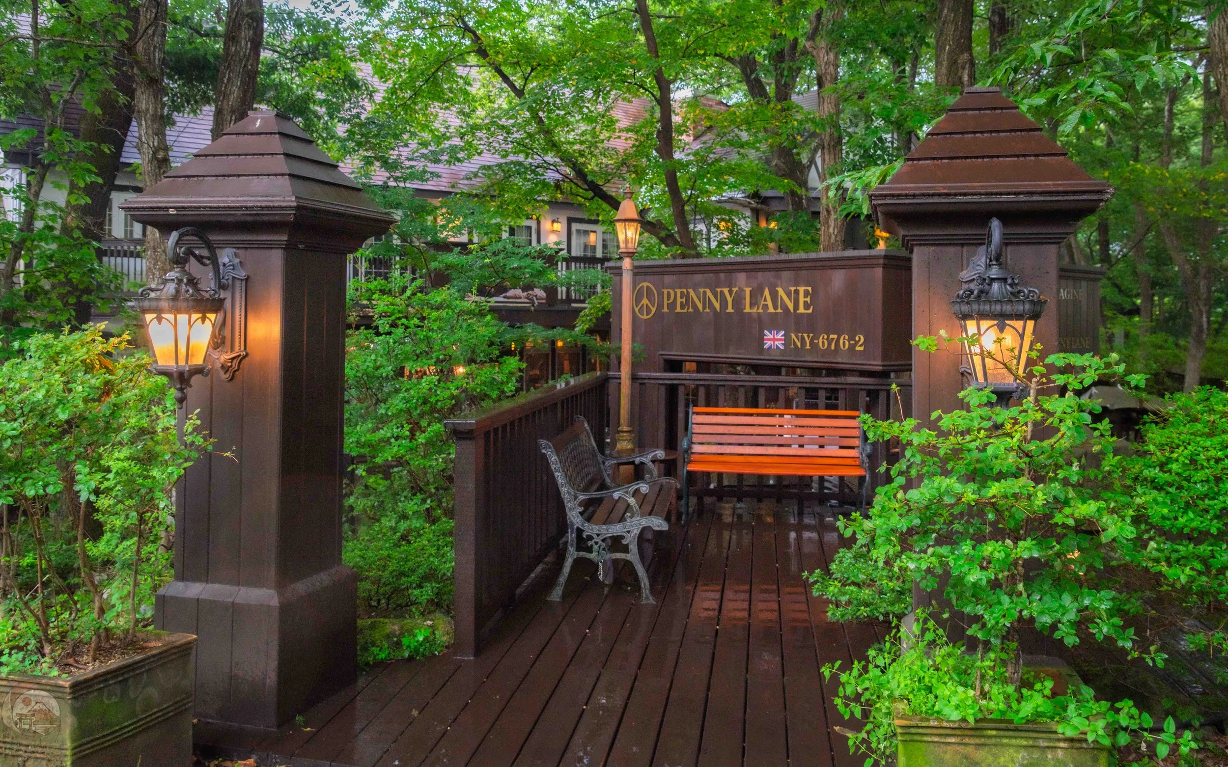 Wooden walkway leading to a bench beneath a sign reading “Penny Lane,” framed by leafy plants and illuminated by lantern-style lights on a rainy day.