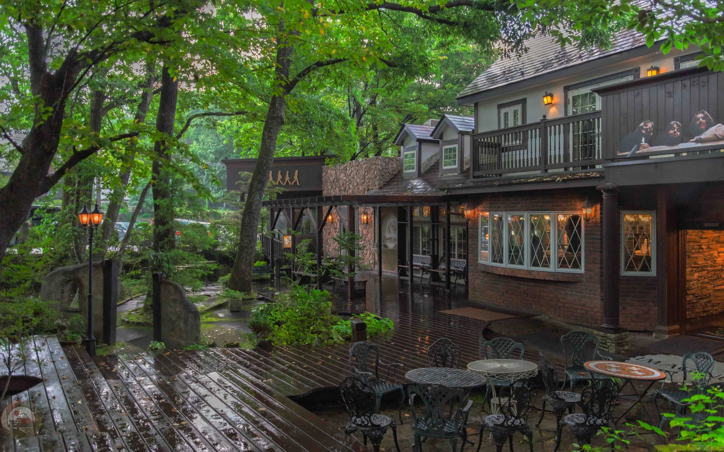Wide view of Penny Lane Bakery with outdoor tables, wet wooden decks, trees, and softly lit windows, showing a quiet garden setting during light rain.