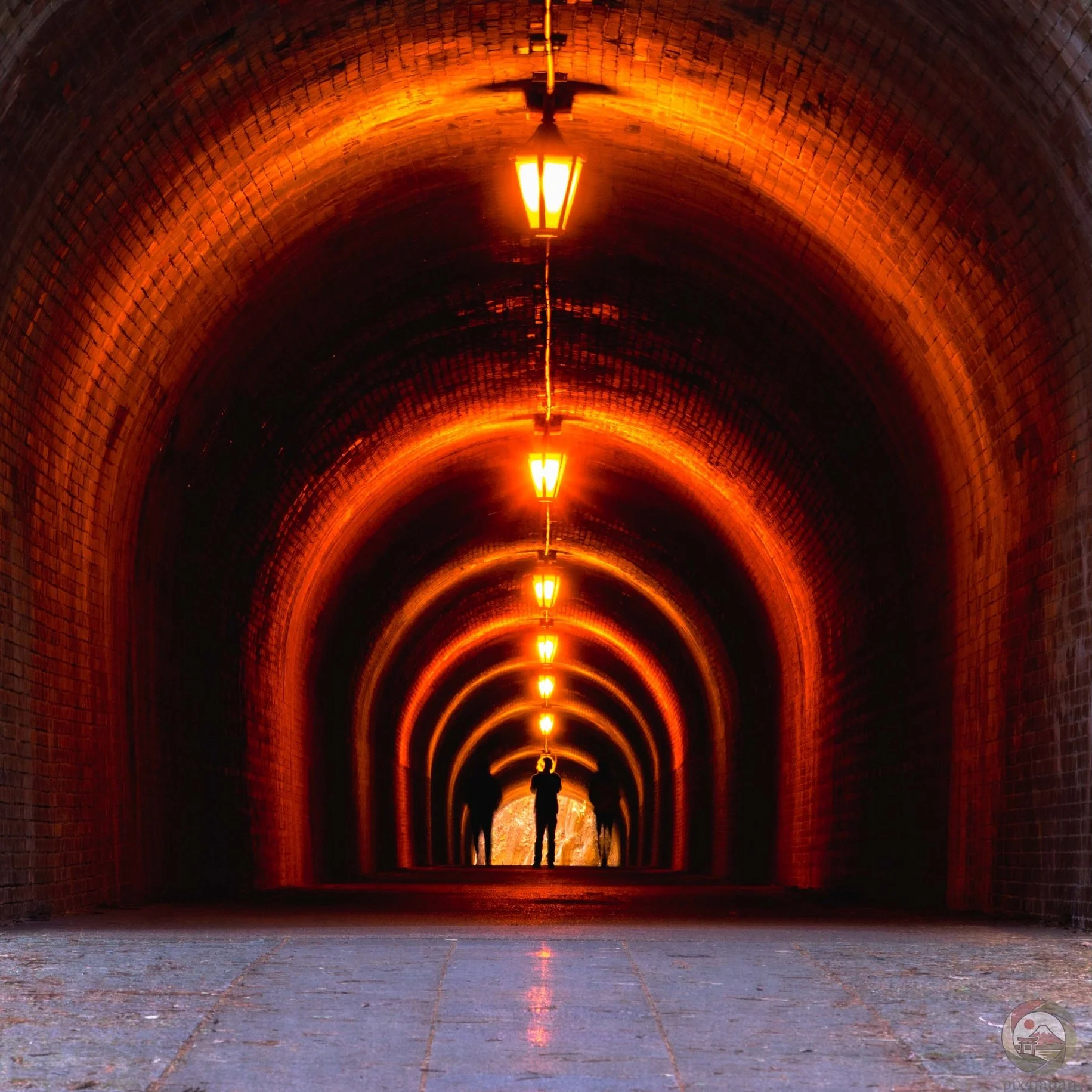Inside a historic brick tunnel, warm orange lamps recede into the distance while three silhouetted people stand near the far exit, where daylight is visible.