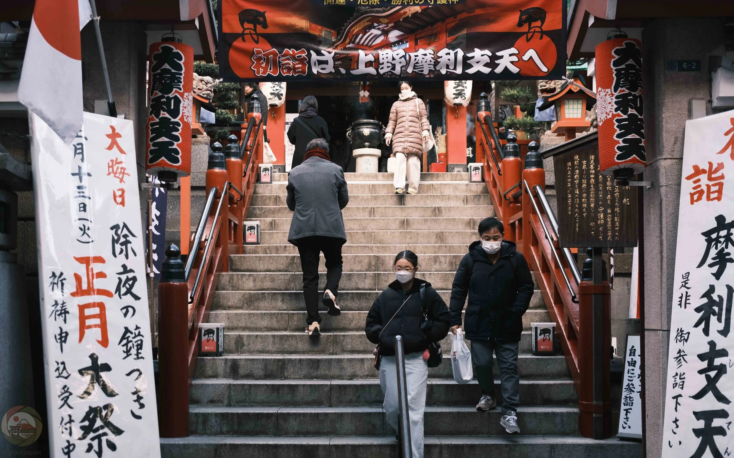 Visitors ascend and descend the stone steps of Marishiten Tokudai-ji, a Buddhist temple in Ueno, Tokyo, during the New Year season, framed by red railings, vertical banners, and hanging paper lanterns.
