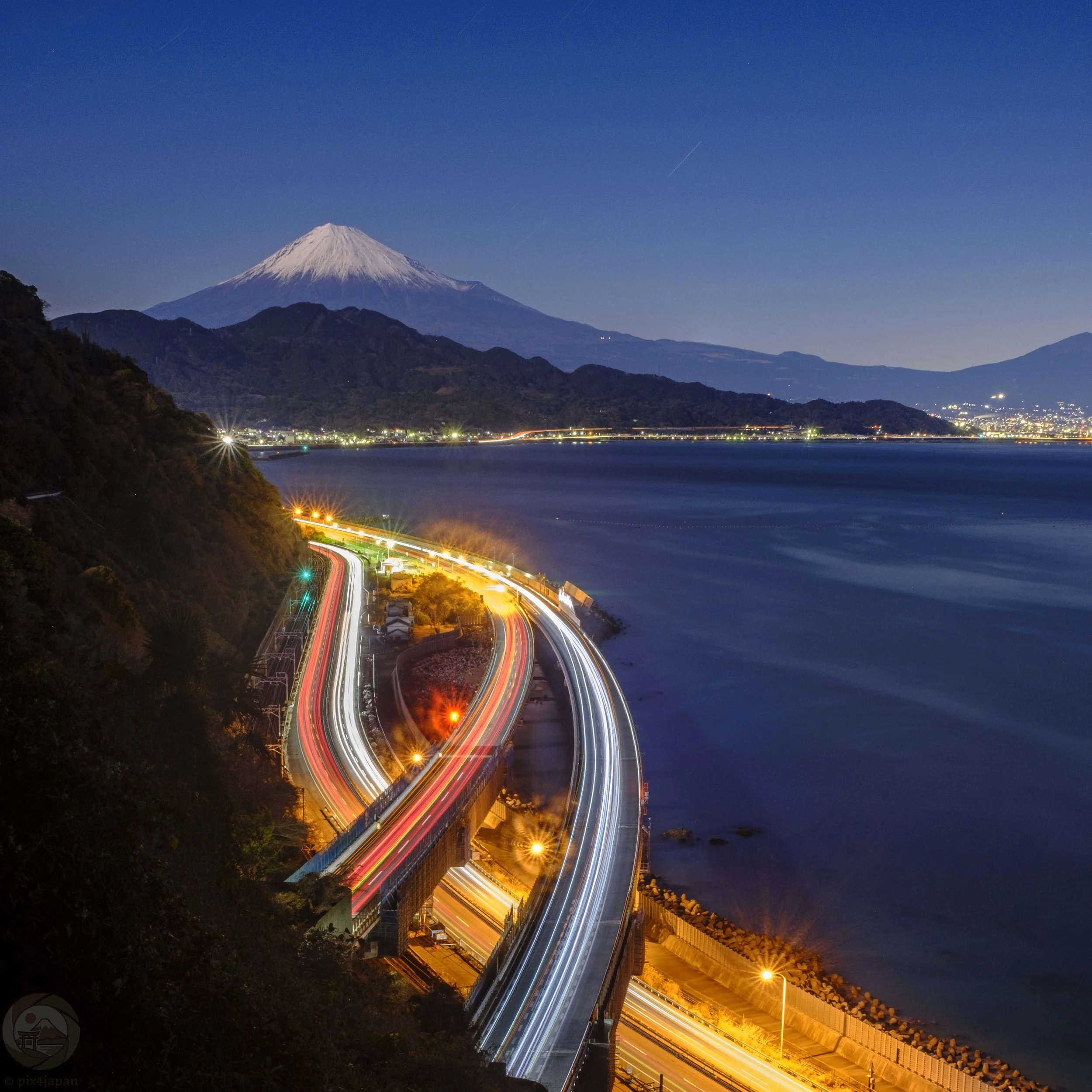 Long-exposure light trails curve along a coastal highway below snow-capped Mount Fuji at blue hour, viewed from Satta-Toge Pass in Shizuoka, Japan.