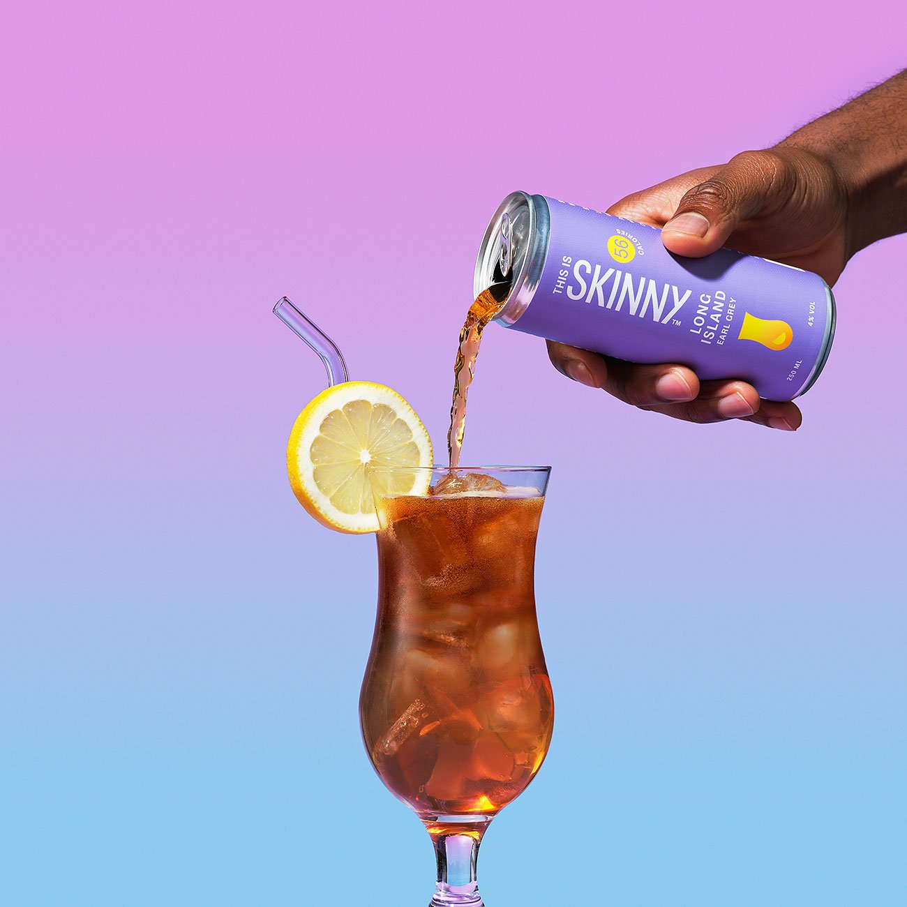 A hand is pouring a This Is Skinny cocktail from a can into a hurricane glass with a lemon garnish.