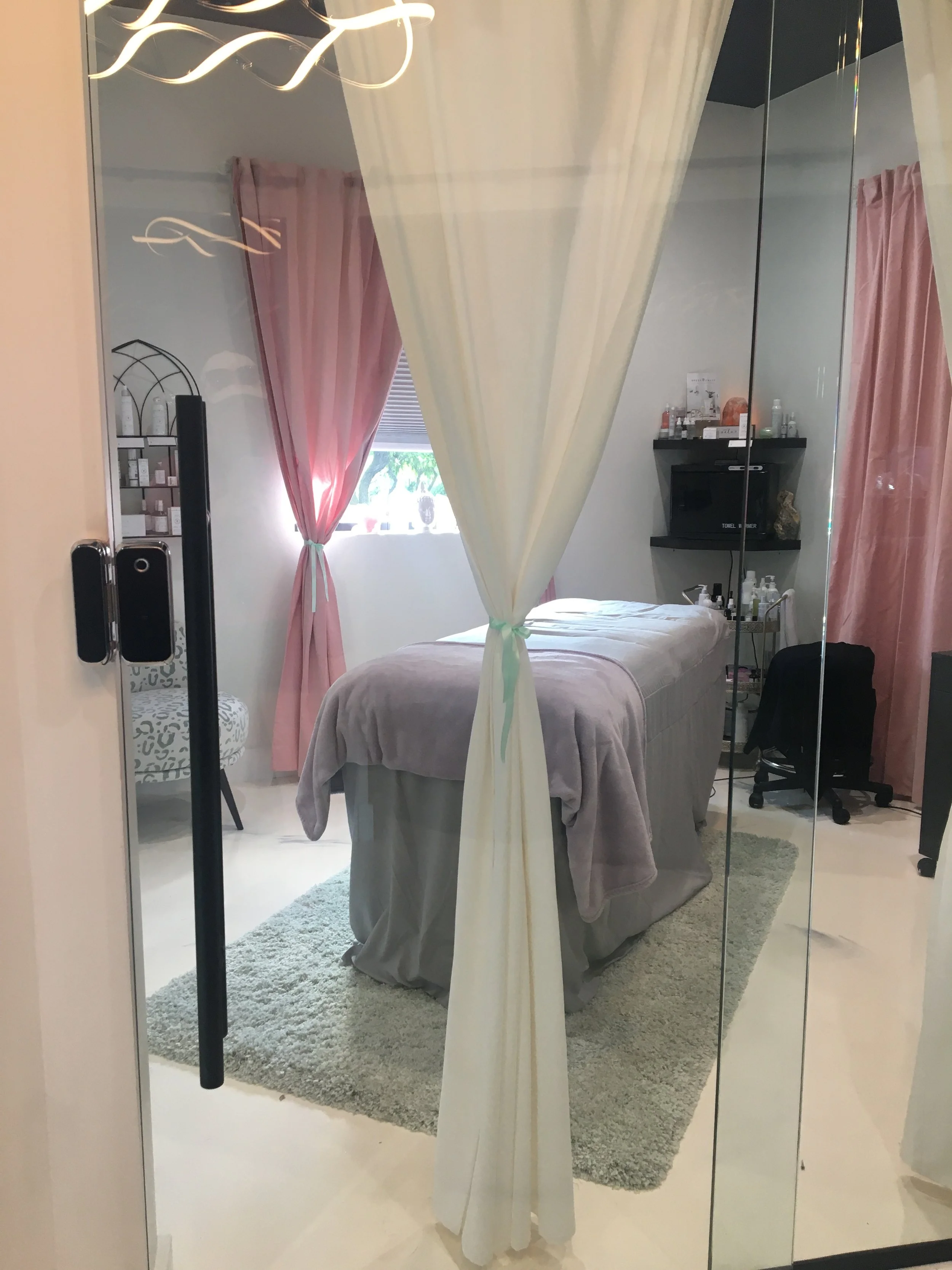 View of a spa or massage room seen through glass doors, with a massage table, pink curtains, and shelves with skincare products.