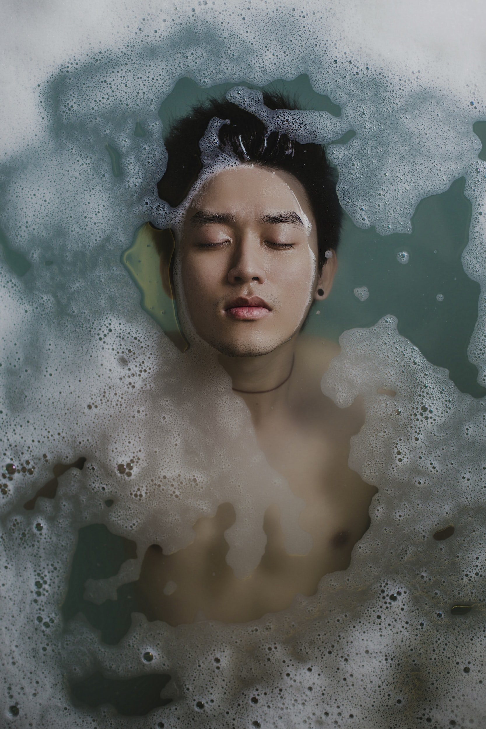 A man with dark hair and closed eyes relaxing in a bath with soap bubbles.