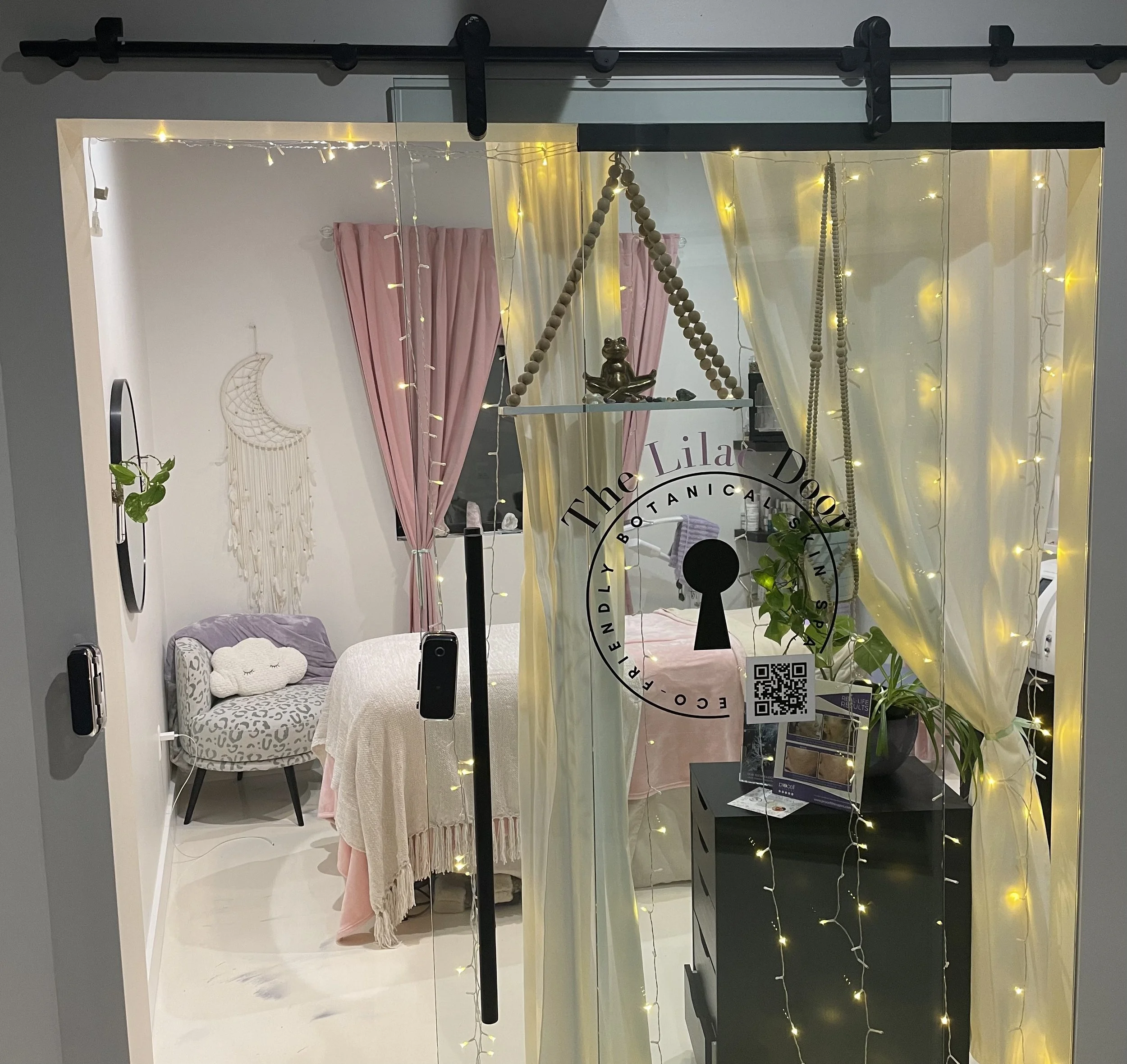 A cozy, aesthetically decorated room viewed through a glass door with fairy lights, pink curtains, a small table with a pink cloth, a gray armchair with a fluffy pillow, and various decorative items including a hanging plaque and a plant.