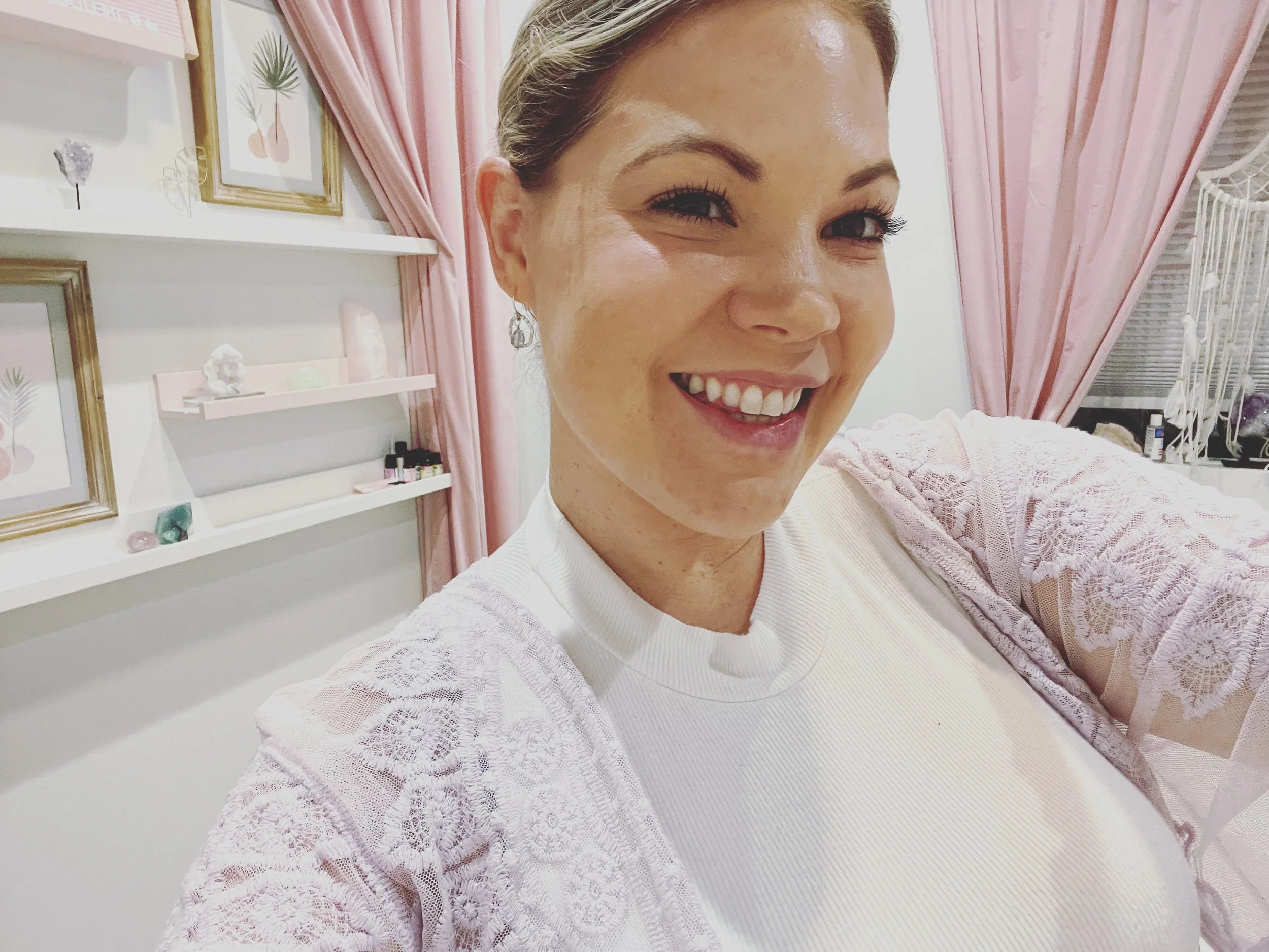 A woman smiling in a room with pink curtains, framed artwork, and decorative items on shelves.