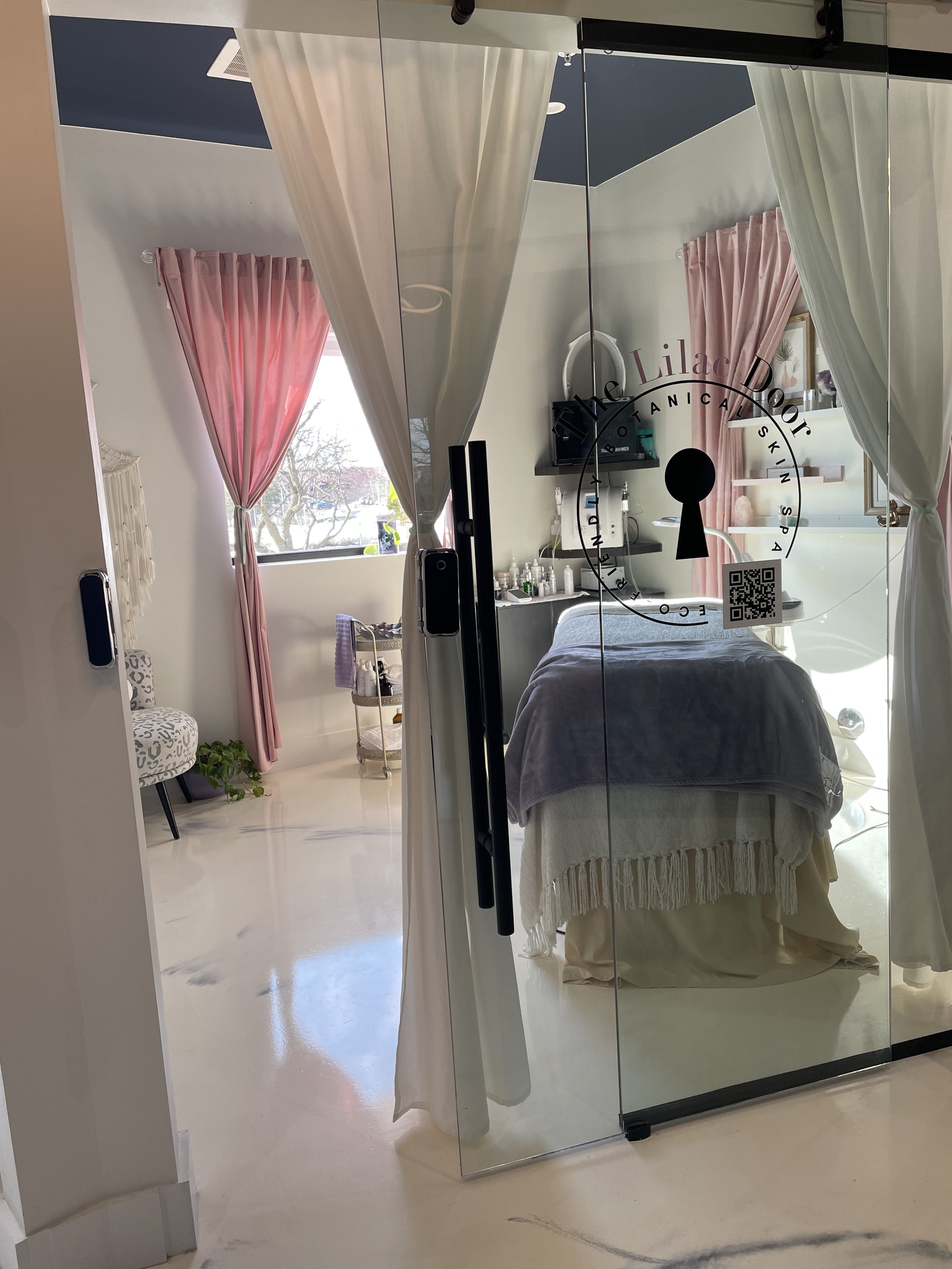 View of a massage therapy room through glass door with the practice's logo, featuring a massage table, pink curtains, a chair, and various supplies in the background.