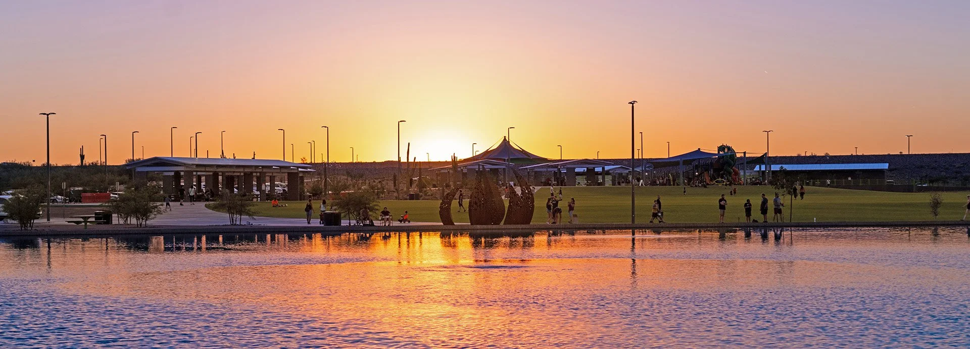Sunset over a park with a body of water in the foreground, a green open field, playground structures, and people walking and playing, with a tent-like pavilion and tall streetlights.