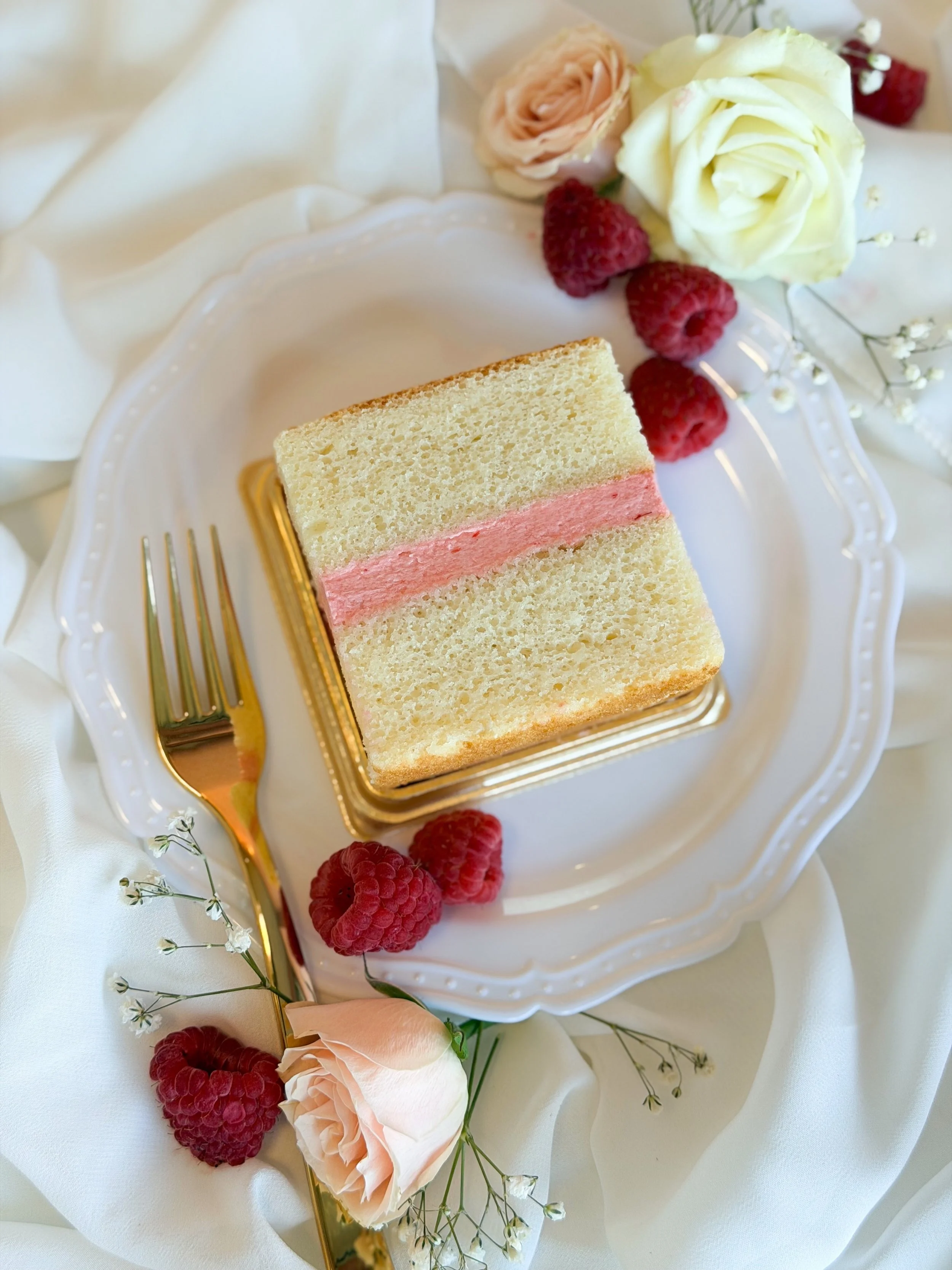 Vanilla cake slice with pink buttercream filling on a china plate with raspberries and fresh flowers