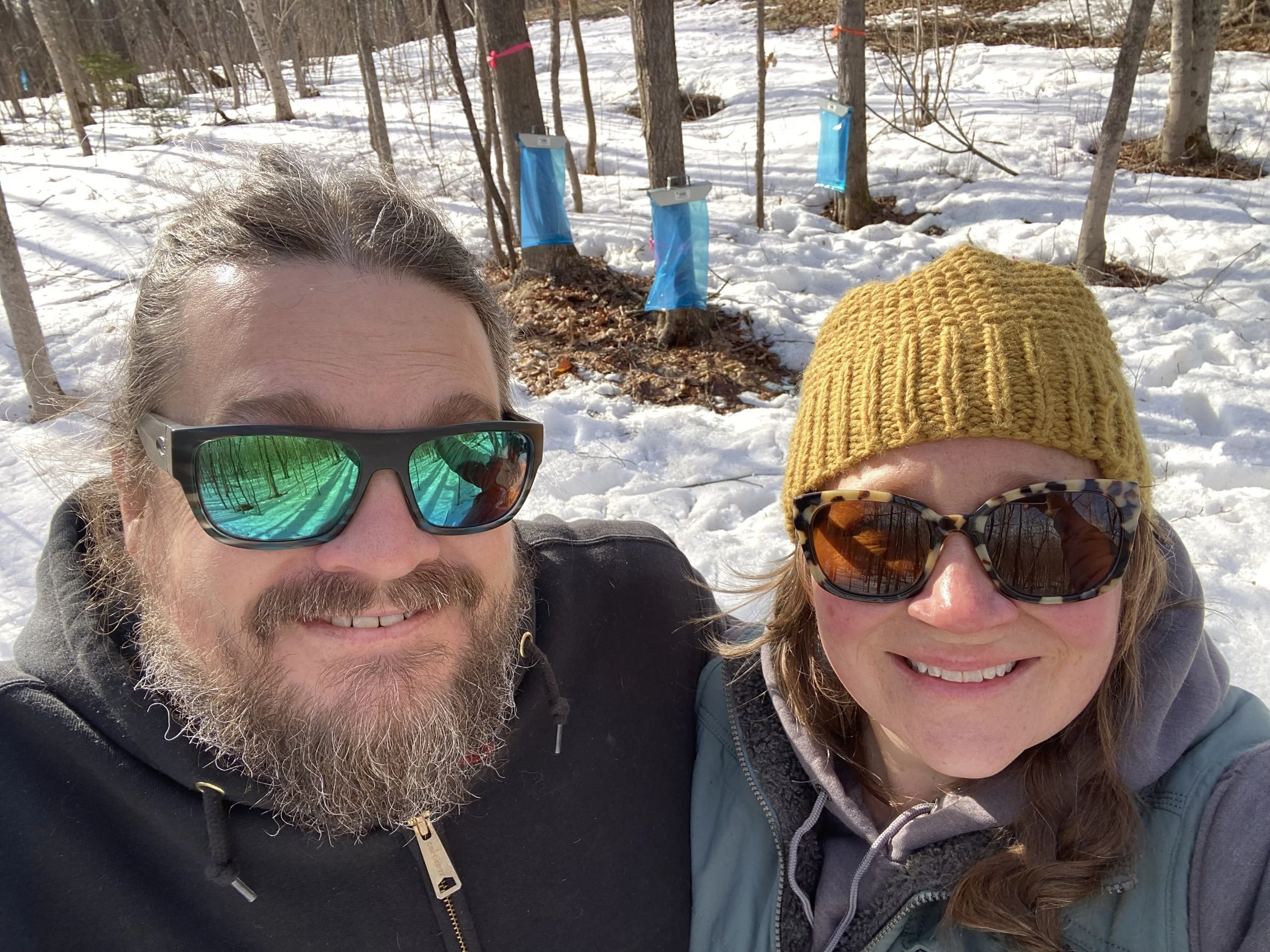 Image of Greg and Megan in their sugarbush, with sap collecting bags on the trees behind them.