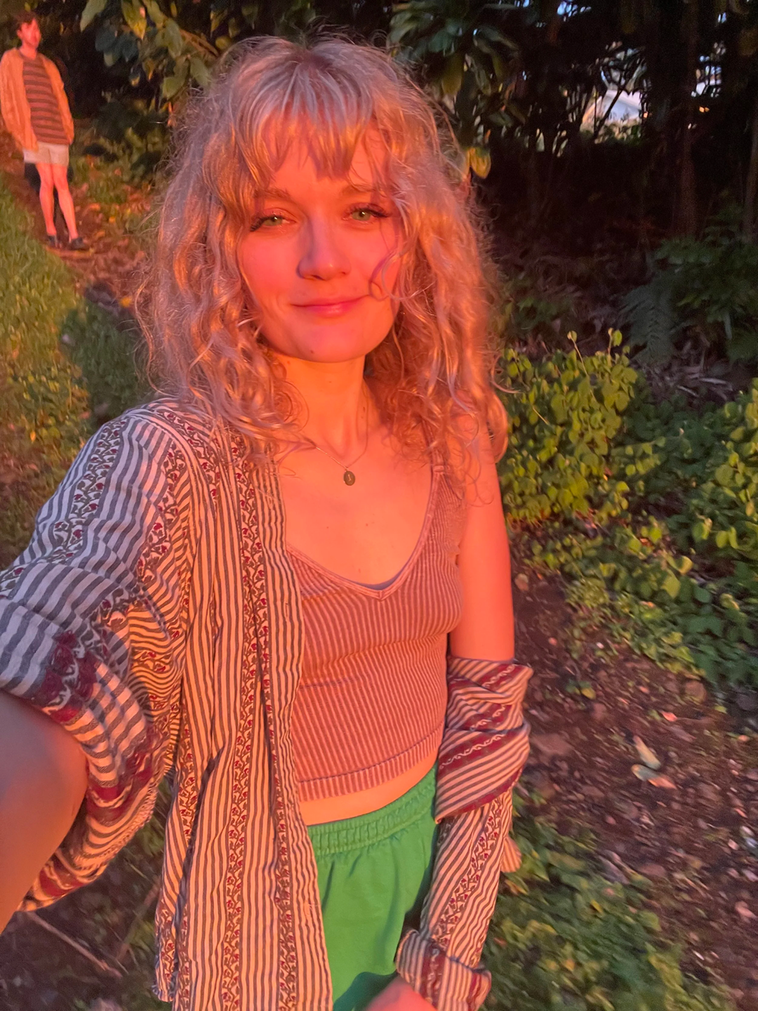 Woman with curly blonde hair smiling outdoors during sunset, wearing a striped crop top and green shorts, with a person in the background standing near bushes.