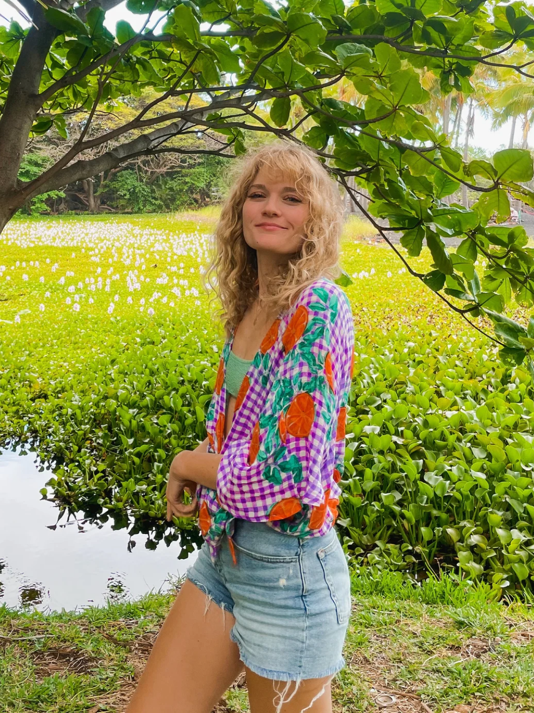 A young woman with curly blonde hair standing outdoors near a pond, under a tree with large green leaves, with a lush green field and white flowers in the background, wearing a colorful patterned blouse and denim shorts.