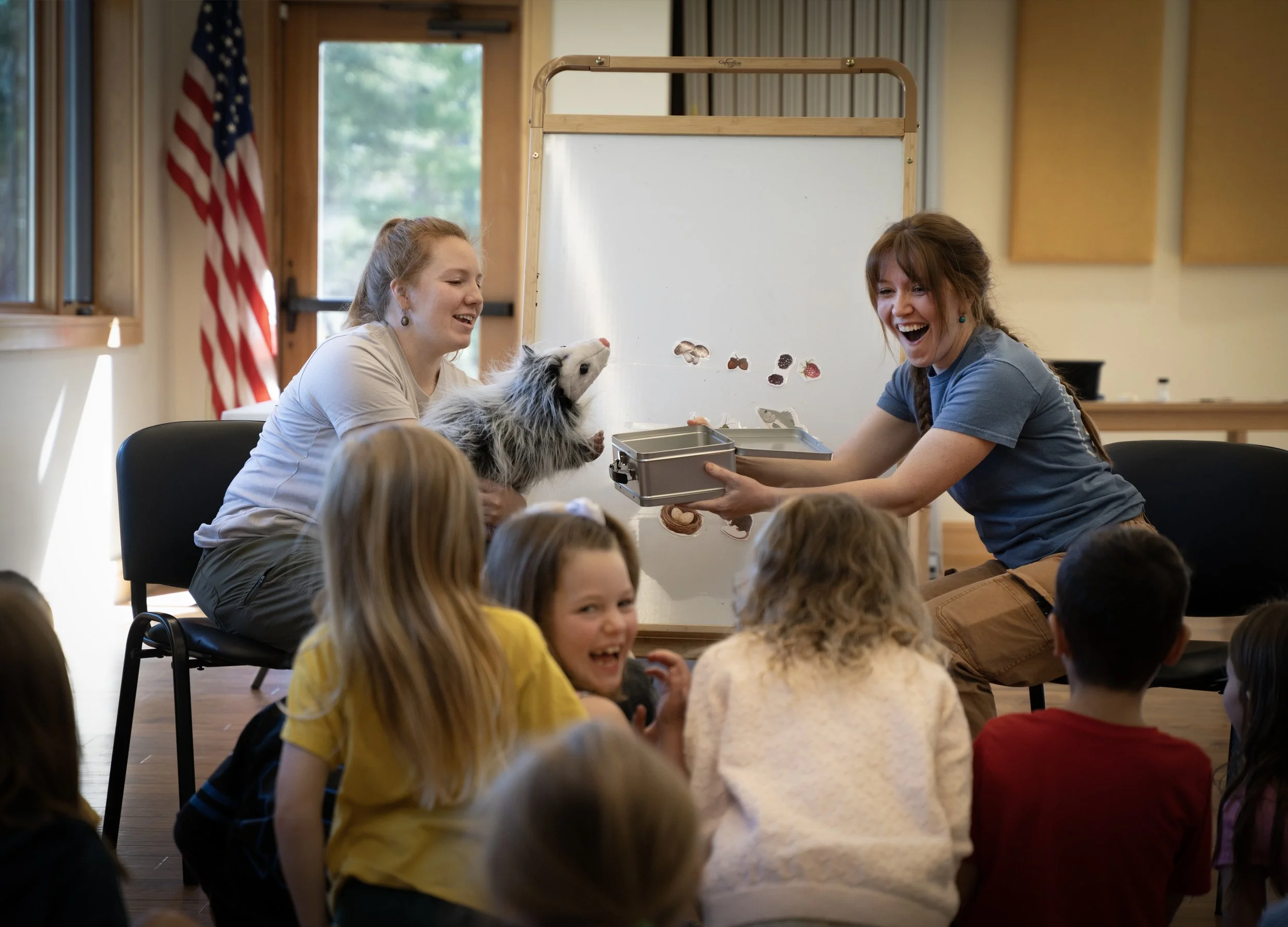 Two women hosting an educational program for kids teaching about what opossums eat. There is a puppet in the hands of one and a lunch box in the hands of the other. The kids are smiling and having a great time.