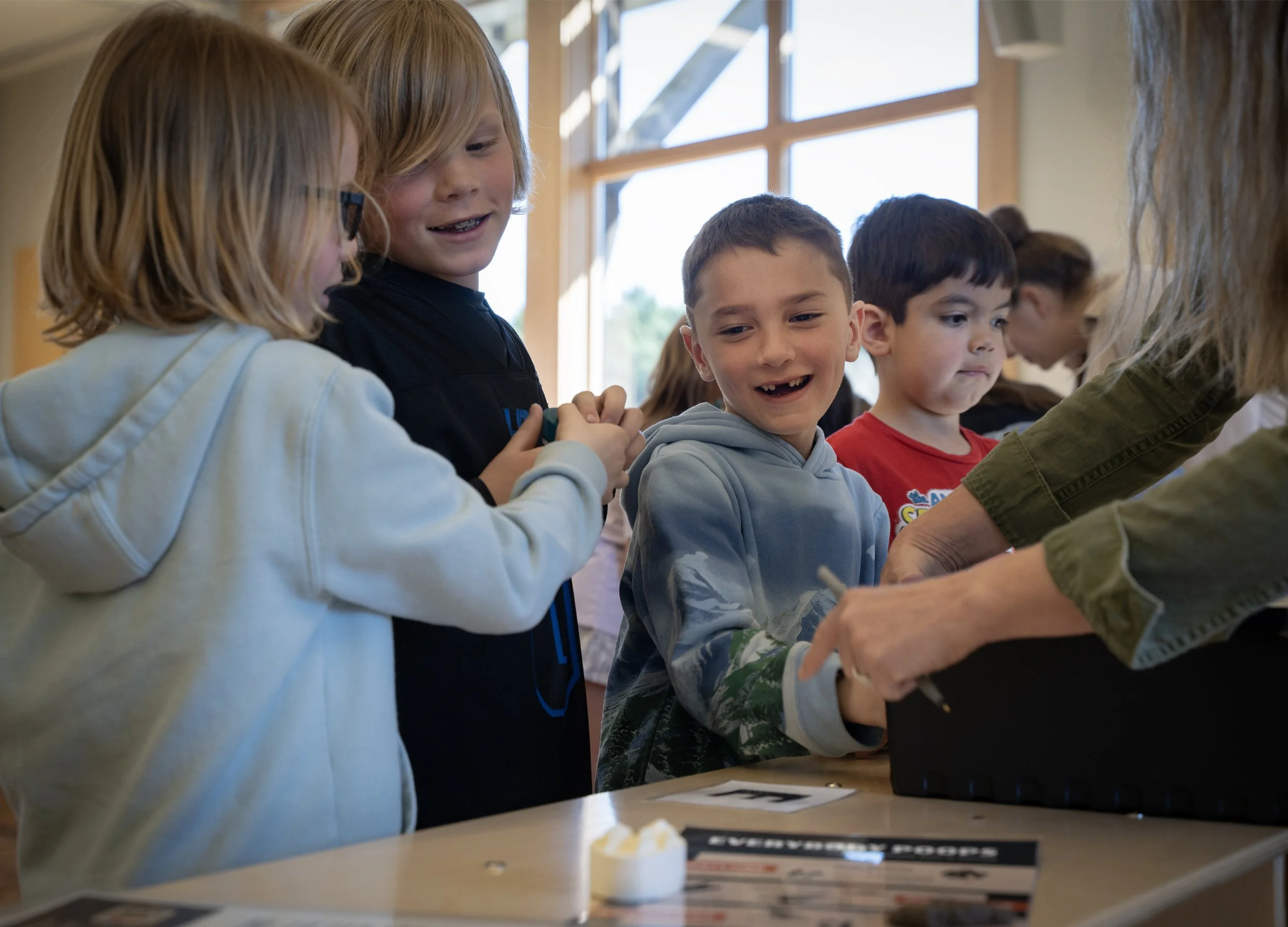 Children gathered in a classroom, engaging in an activity at a table, with a window in the background letting in natural light.