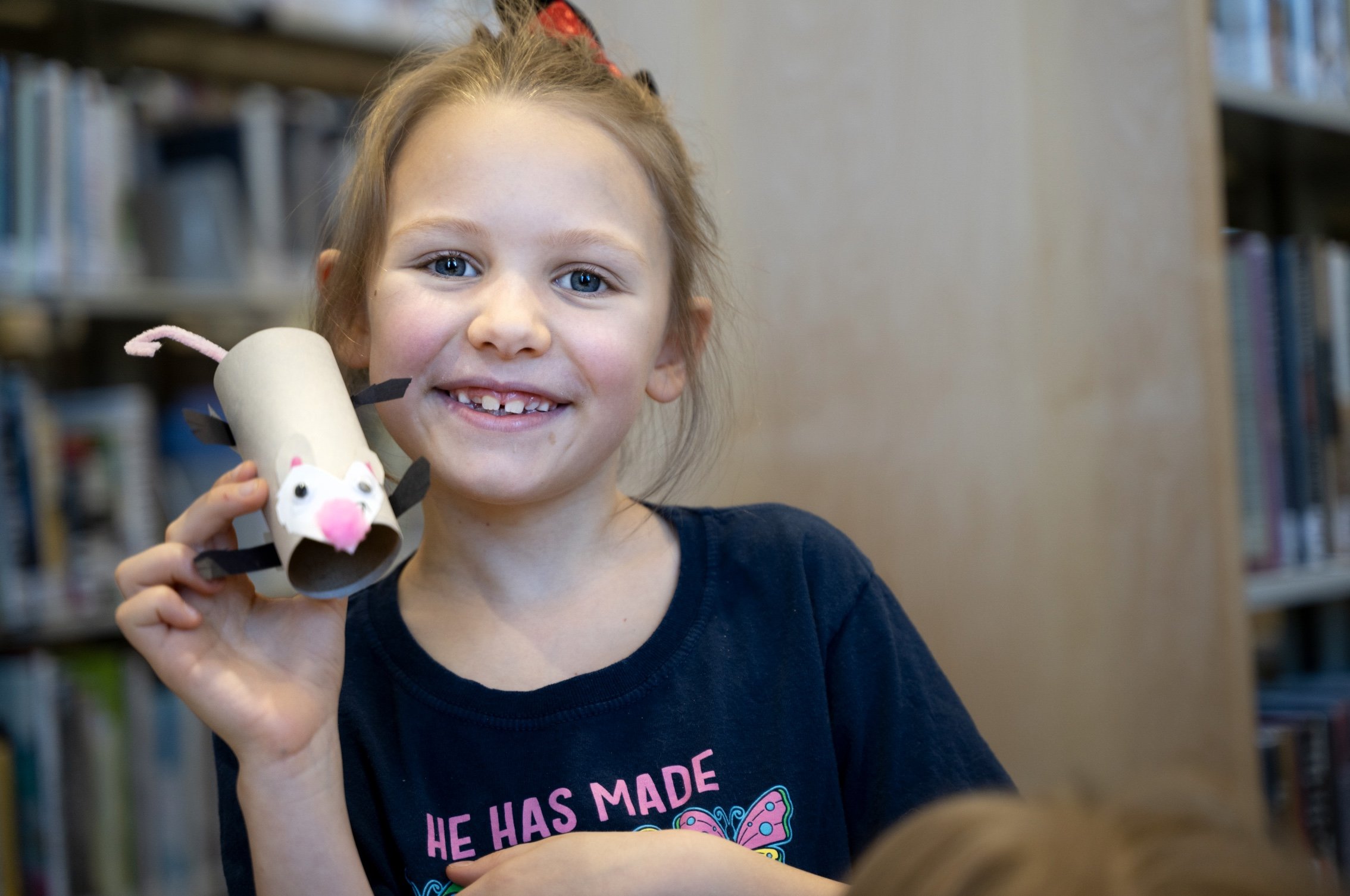 A young girl with blue eyes and a big smile holding a handmade craft opossum made from a toilet paper roll, in a library.