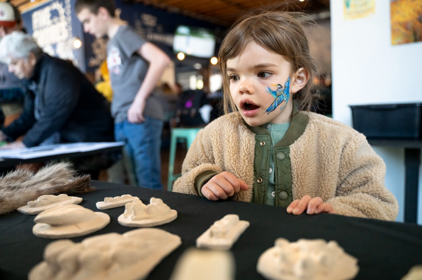 Outreach education table A young girl with a painted blue and yellow bird on her face, wearing a beige fluffy jacket, sitting at a table with various animal-shaped cookies, at an indoor event or market.