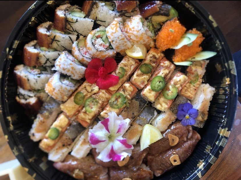 Assorted sushi platter with colorful rolls, grilled skewers, spicy peppers, lemon wedges, and edible flowers