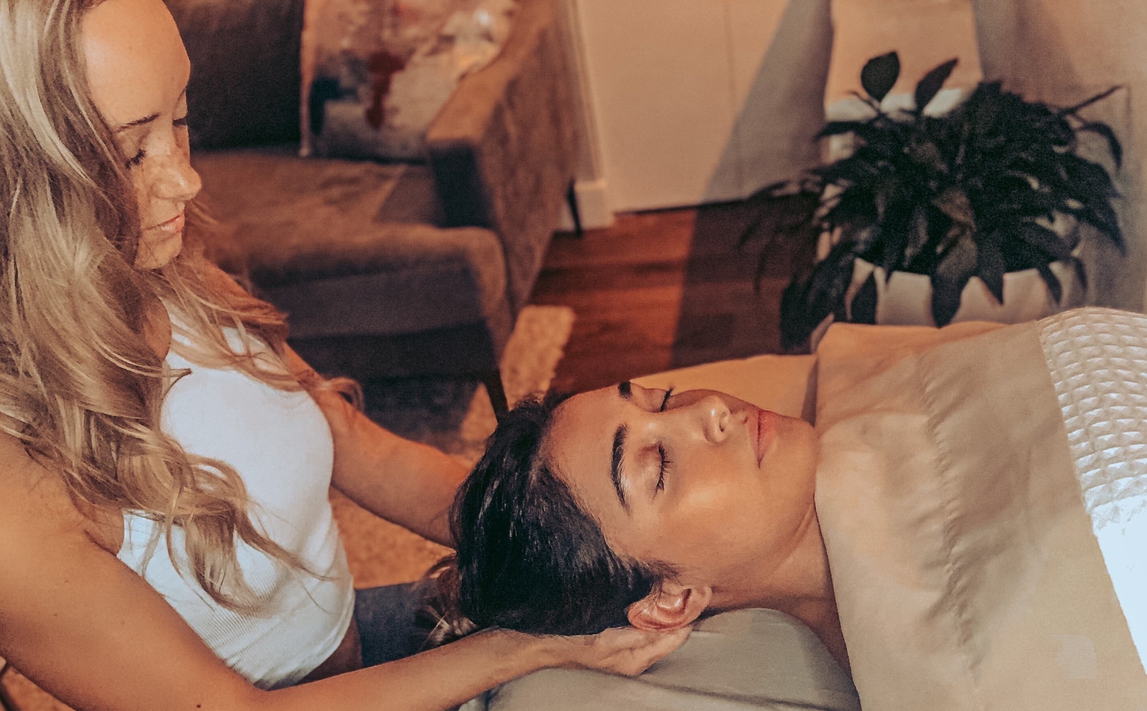 A woman with dark hair and closed eyes resting her head on a pillow, a blonde woman with wavy hair and a white shirt leaning over her, holding her face gently in a cozy indoor setting with a brown couch, wooden furniture, and a potted plant in the background.