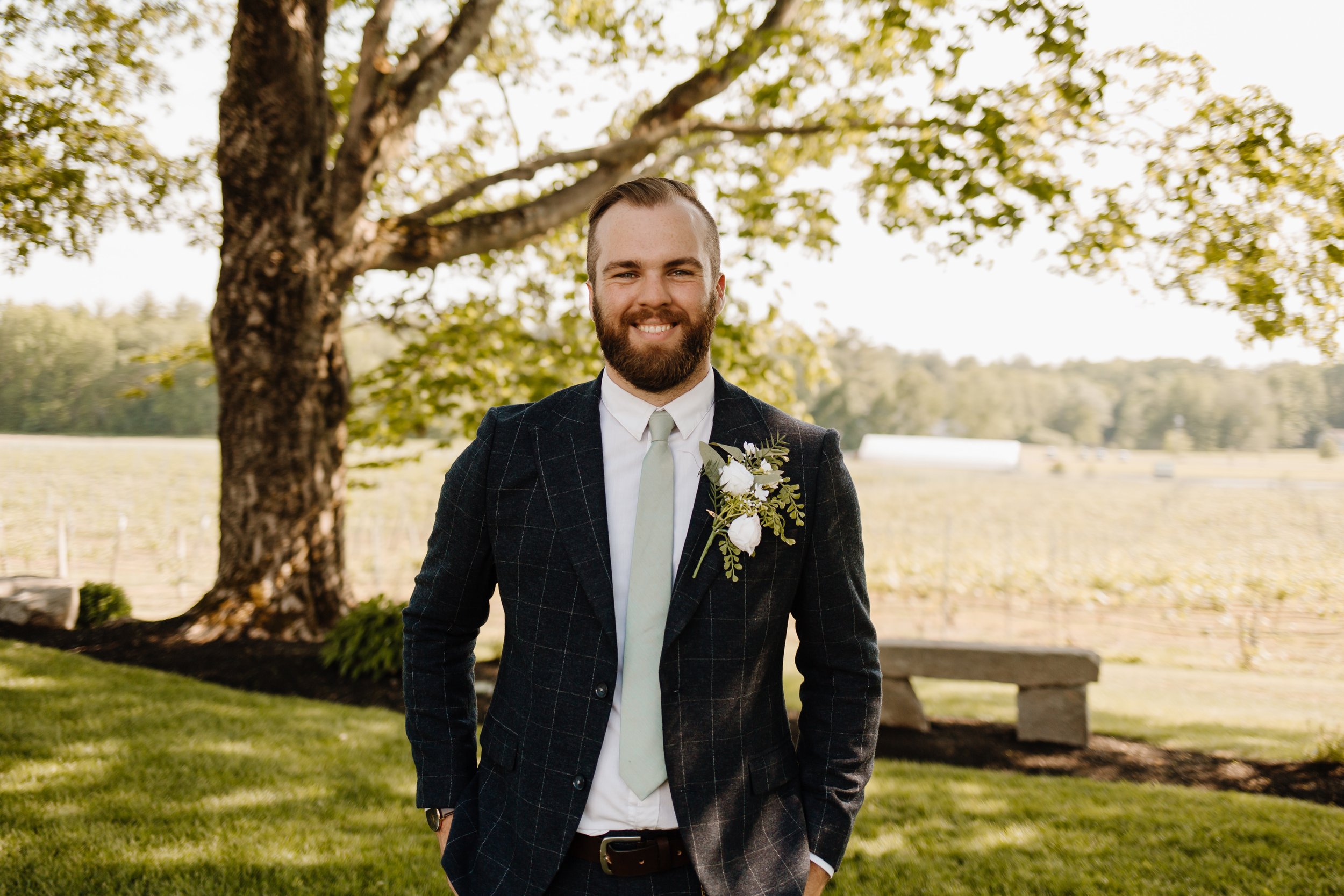 A man in a dark checkered suit with a white shirt and pale green tie, standing outdoors under a large tree, smiling at the camera, with a field and hay bales in the background.