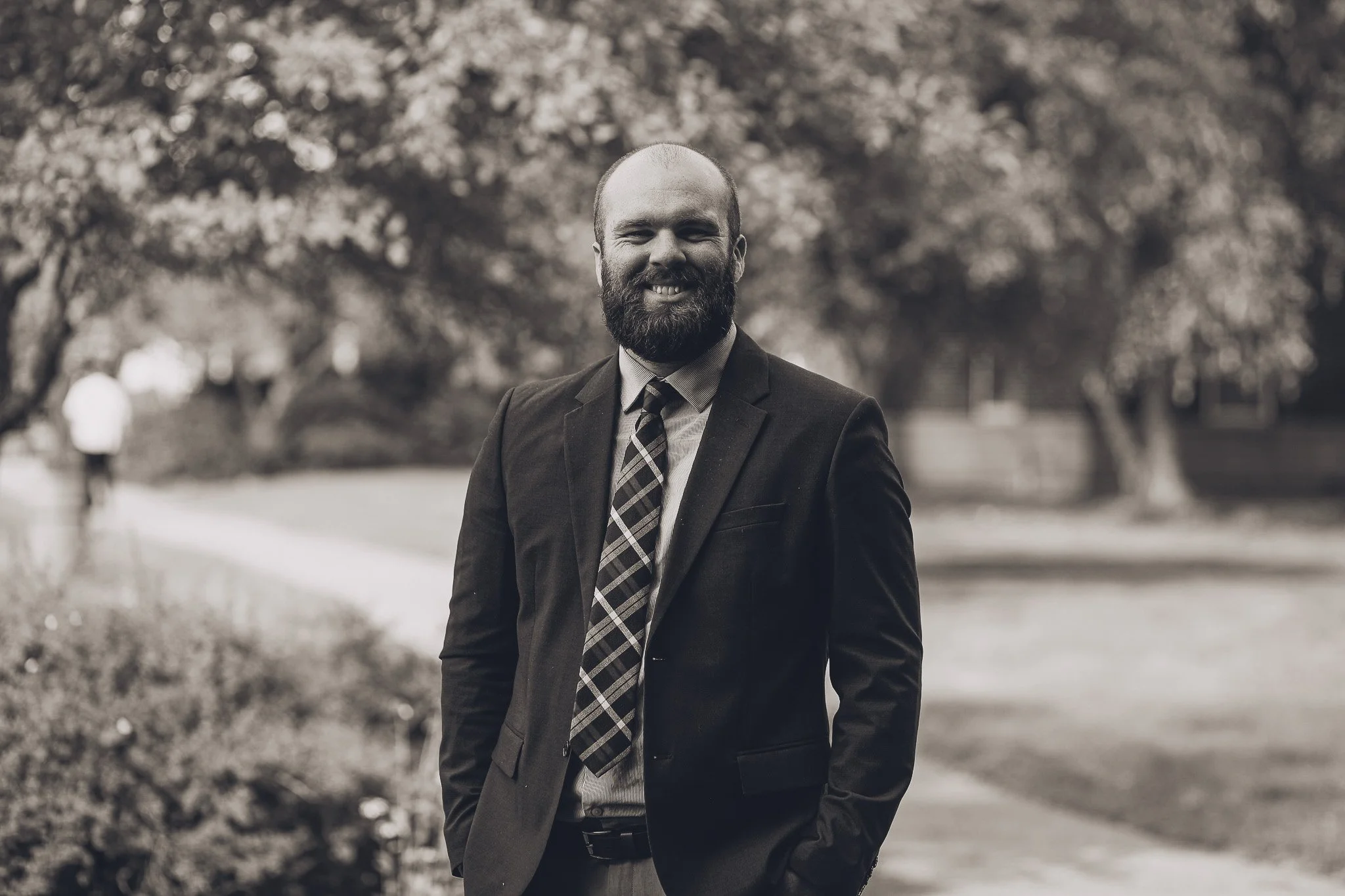 A man with a beard and bald head smiling outdoors, wearing a dark suit and a patterned tie, standing on a park path with trees and blurred figures in the background.
