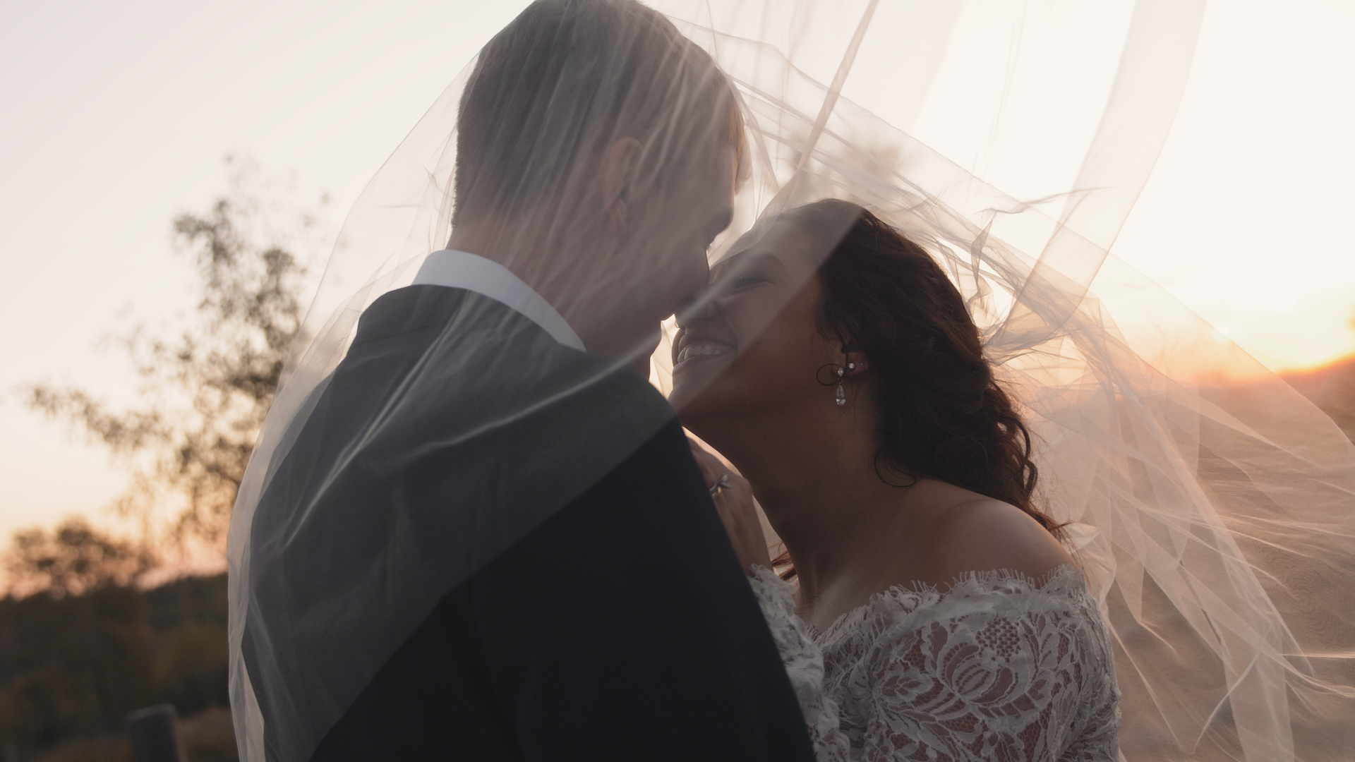 A bride and groom share an intimate moment under a wedding veil during sunset, with trees in the background.