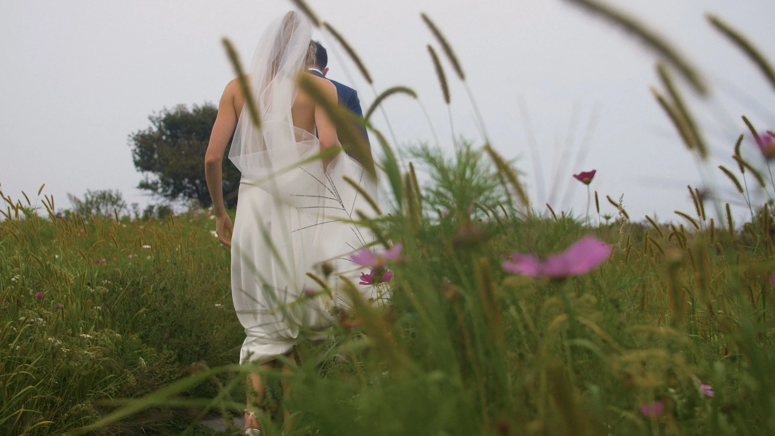 A bride in a wedding dress and veil walking through a grassy field with pink flowers, viewed from below and behind, with a tree and cloudy sky in the background.