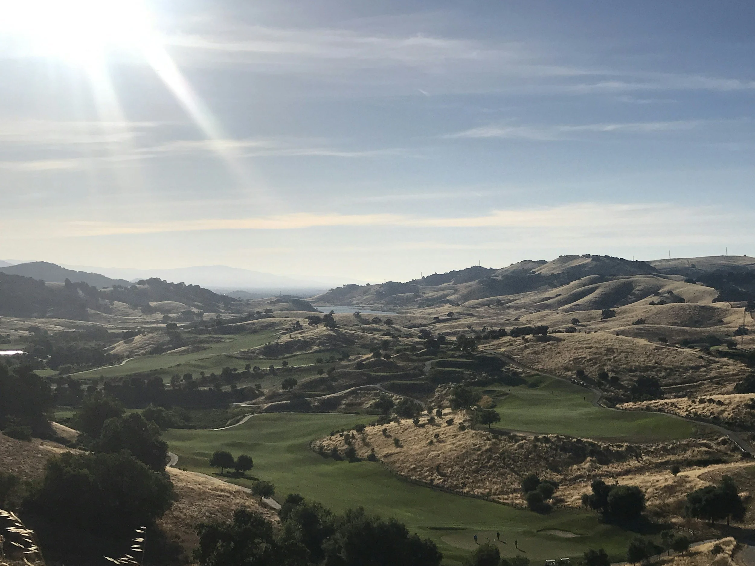 Sunlit landscape of rolling hills with green patches and scattered trees, under a partly cloudy sky.