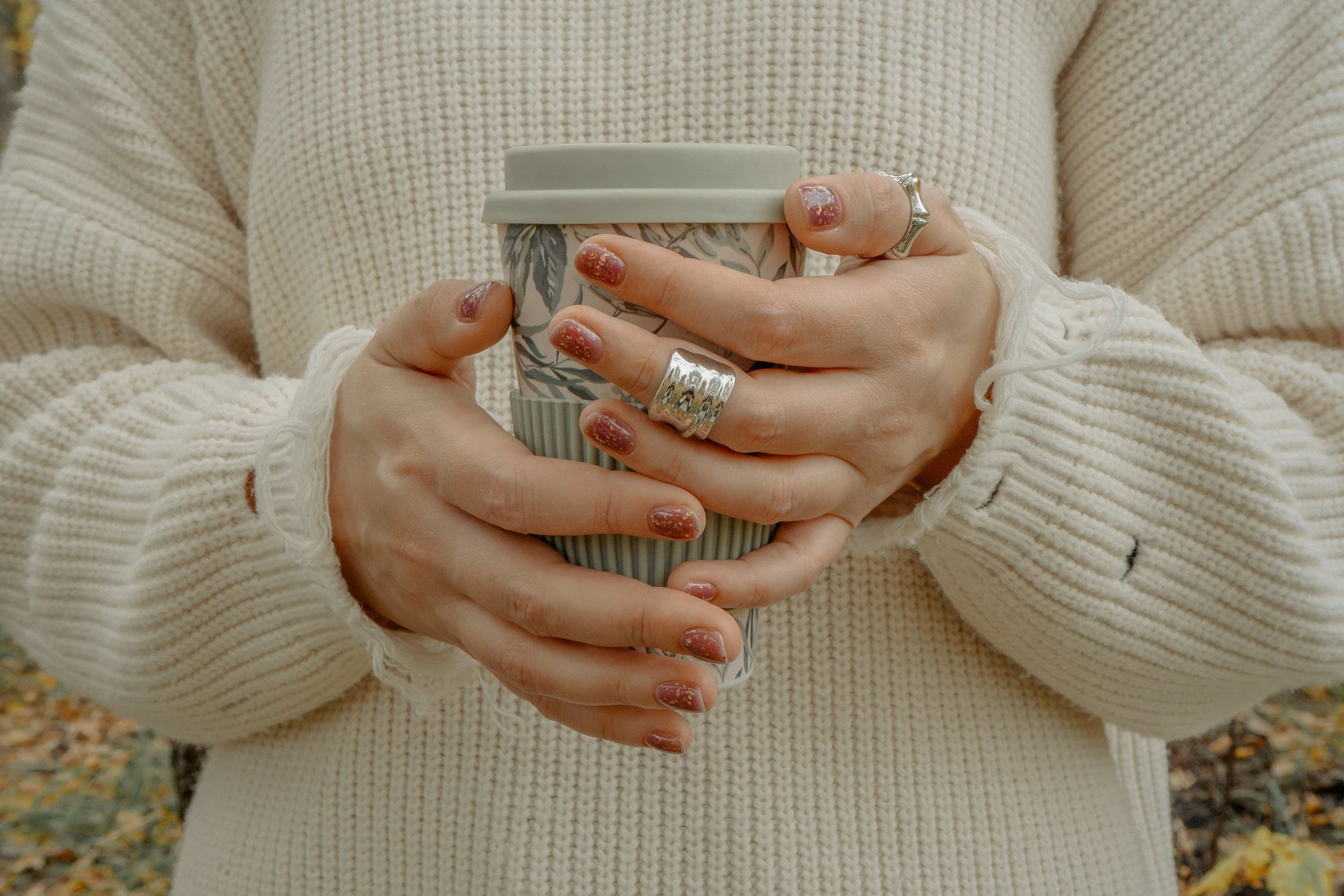 Person holding a to-go coffee cup with both hands, wearing a cream-colored sweater and several rings.