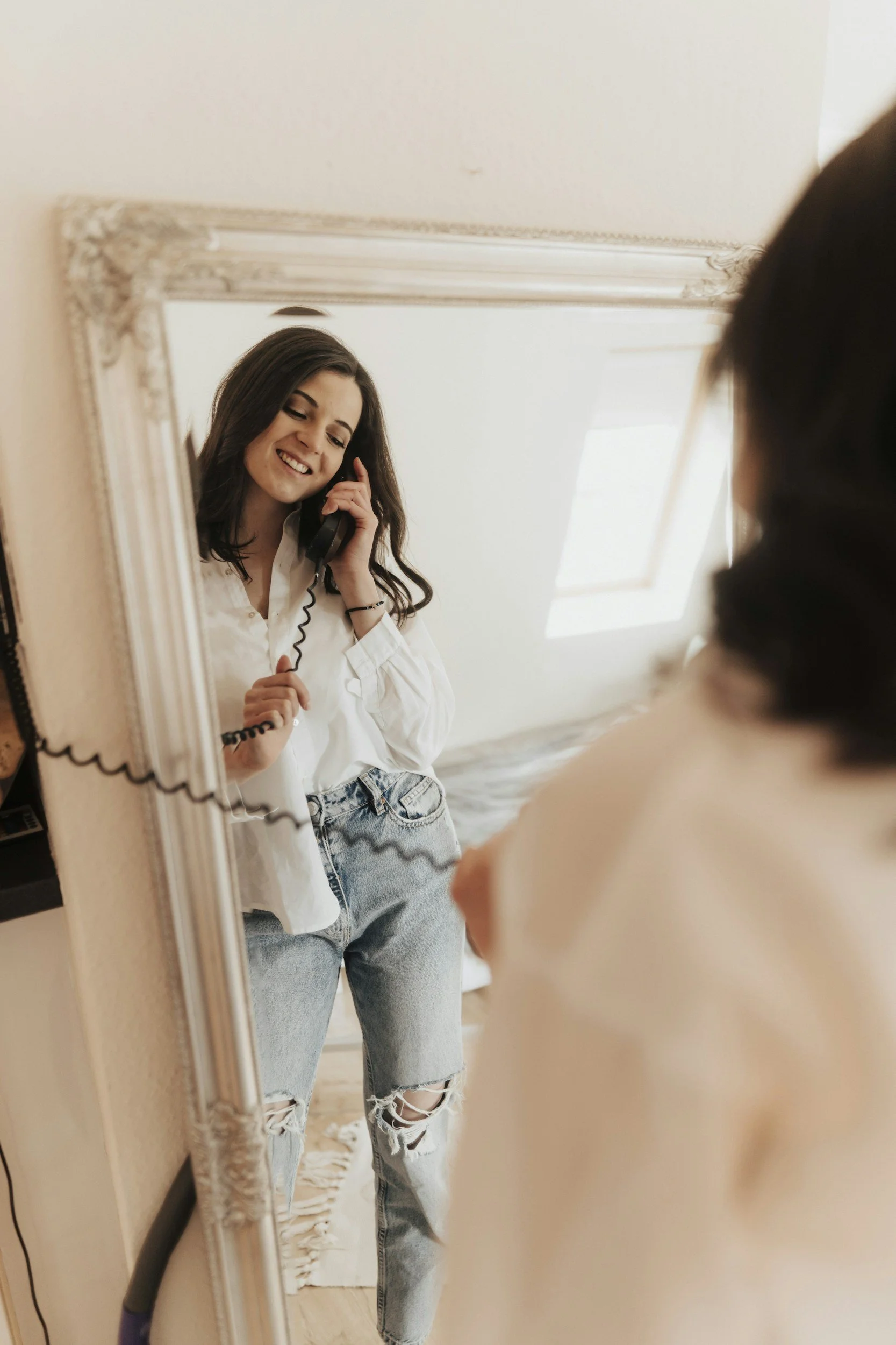 Young woman with long dark hair, wearing a white shirt and ripped jeans, smiling while talking on a phone, taking a selfie in a mirror.