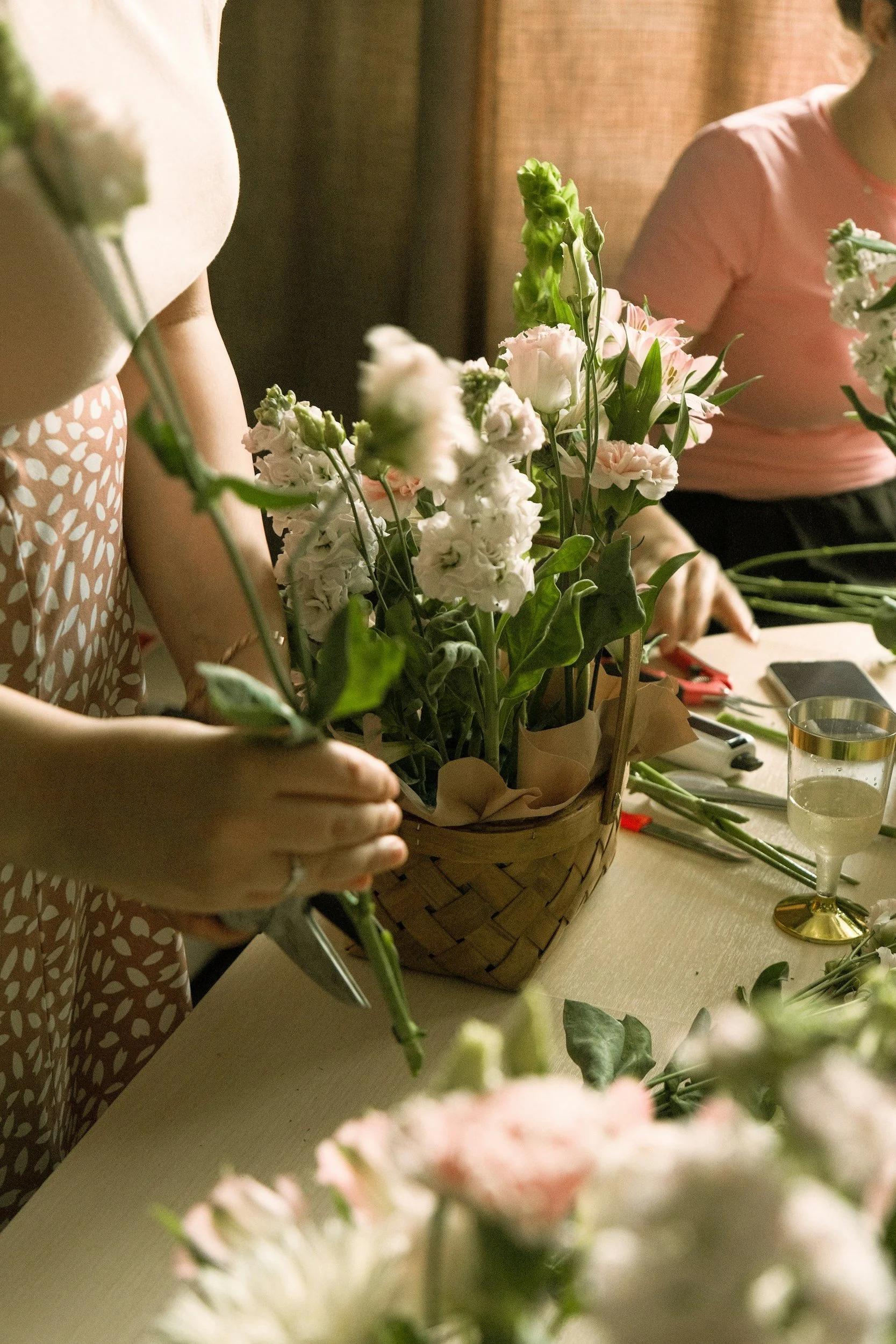 Two women are arranging pink and white flowers in a basket on a table. One woman is wearing a pink top, and the other a patterned dress. There are tools and a glass of water on the table, and a brown curtain in the background.