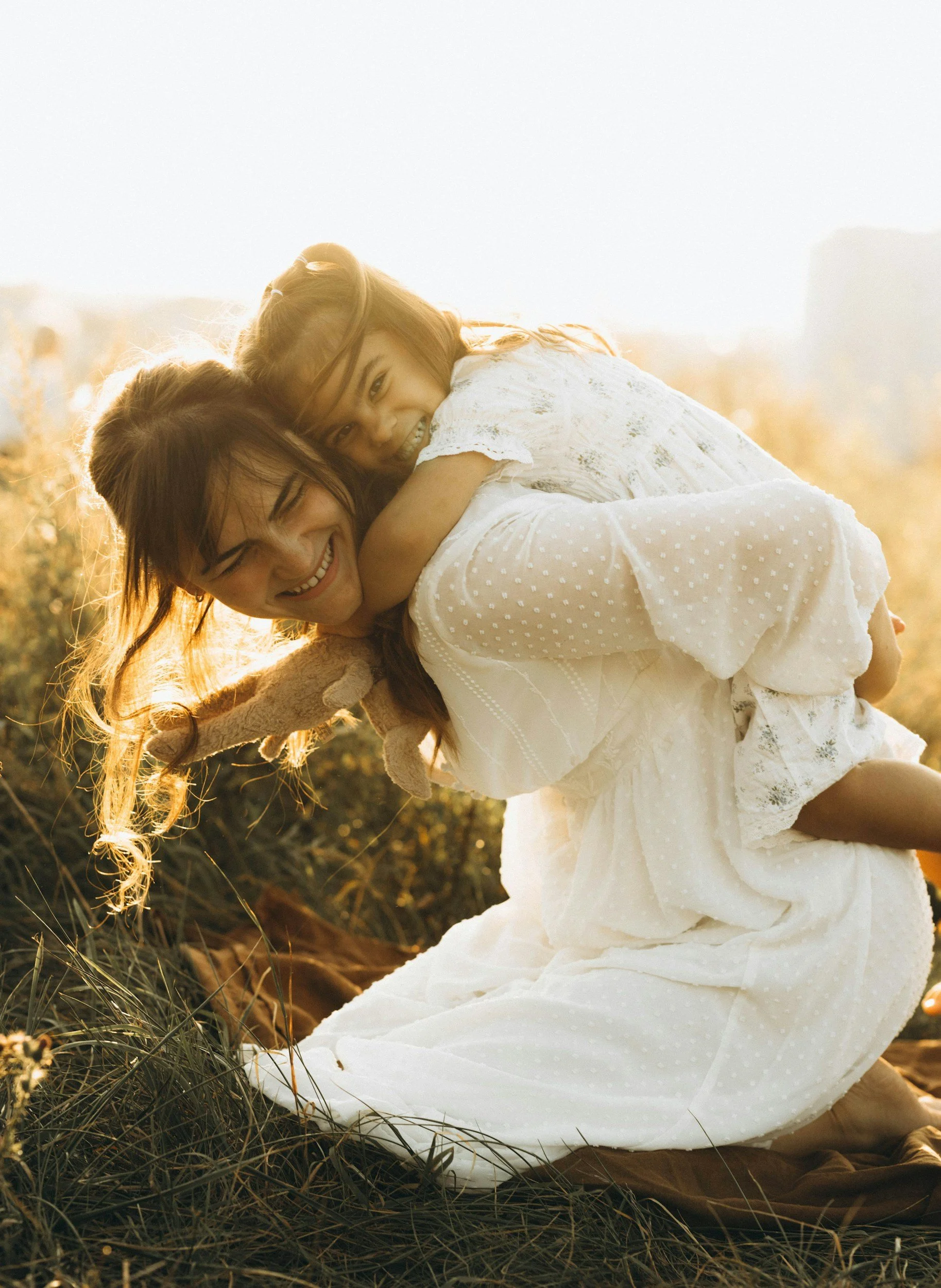 A woman carrying a young girl on her back outdoors during sunset, both smiling warmly, surrounded by grass and warm sunlight.