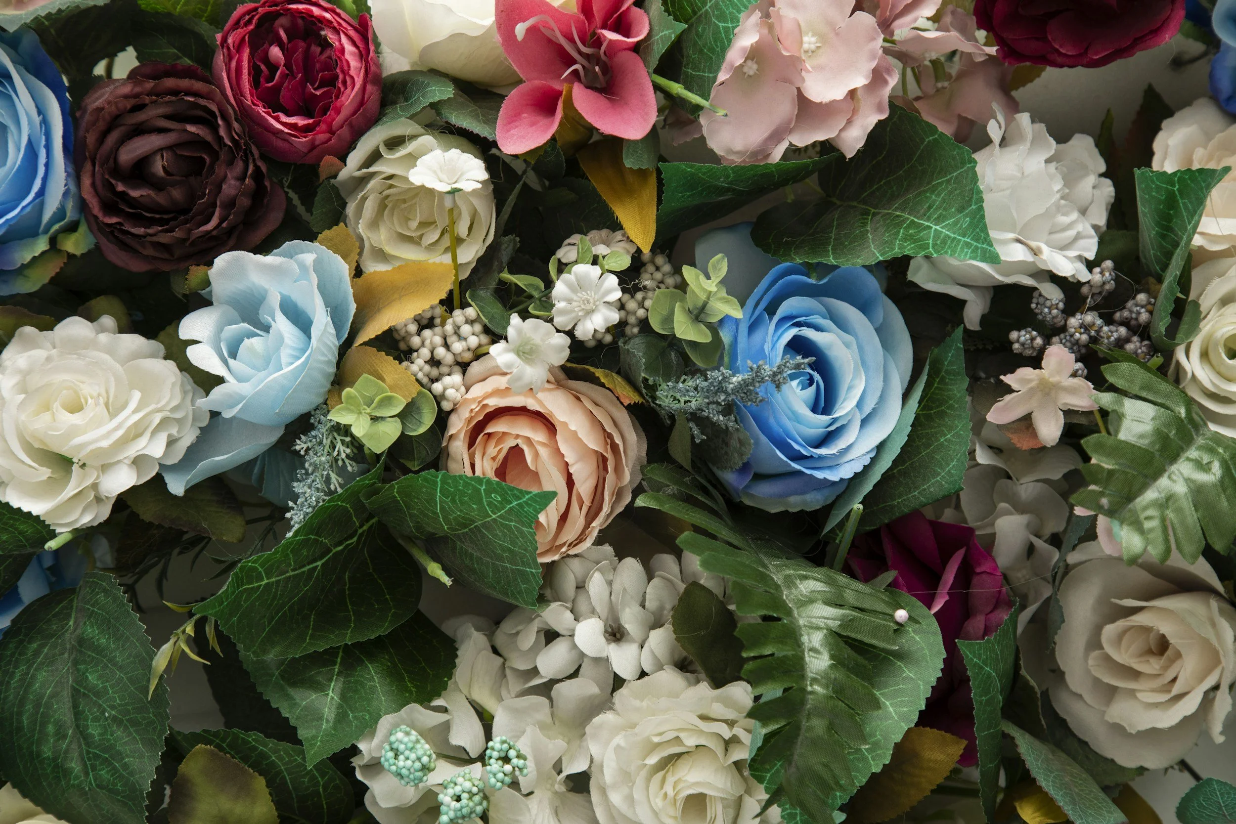 A collection of colorful artificial flowers, including roses, hydrangeas, and other blossoms, with green leaves and small decorative berry accents.