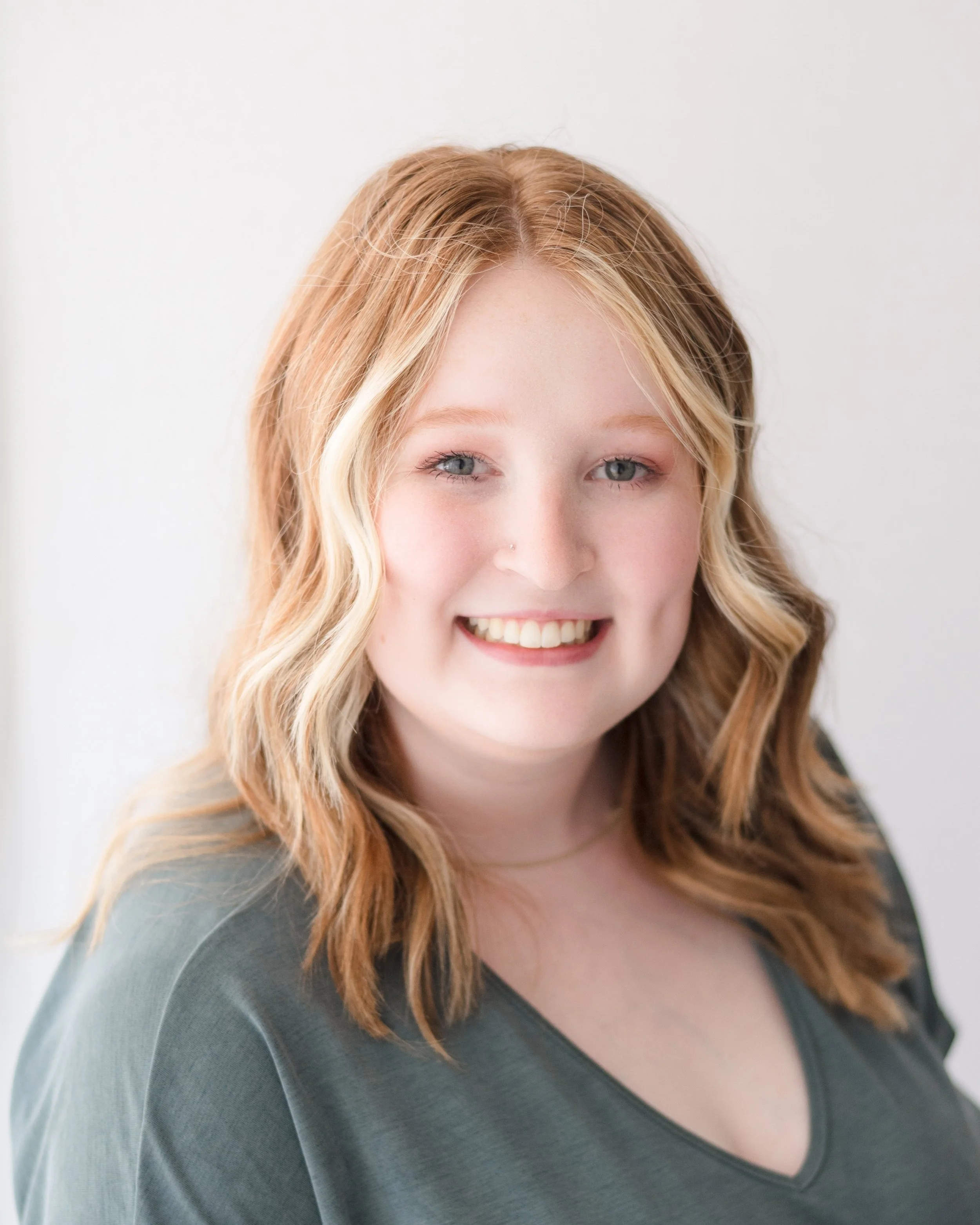 A young woman with wavy, shoulder-length blonde hair, light skin, and blue eyes smiling, wearing a dark gray top, in front of a plain white background.