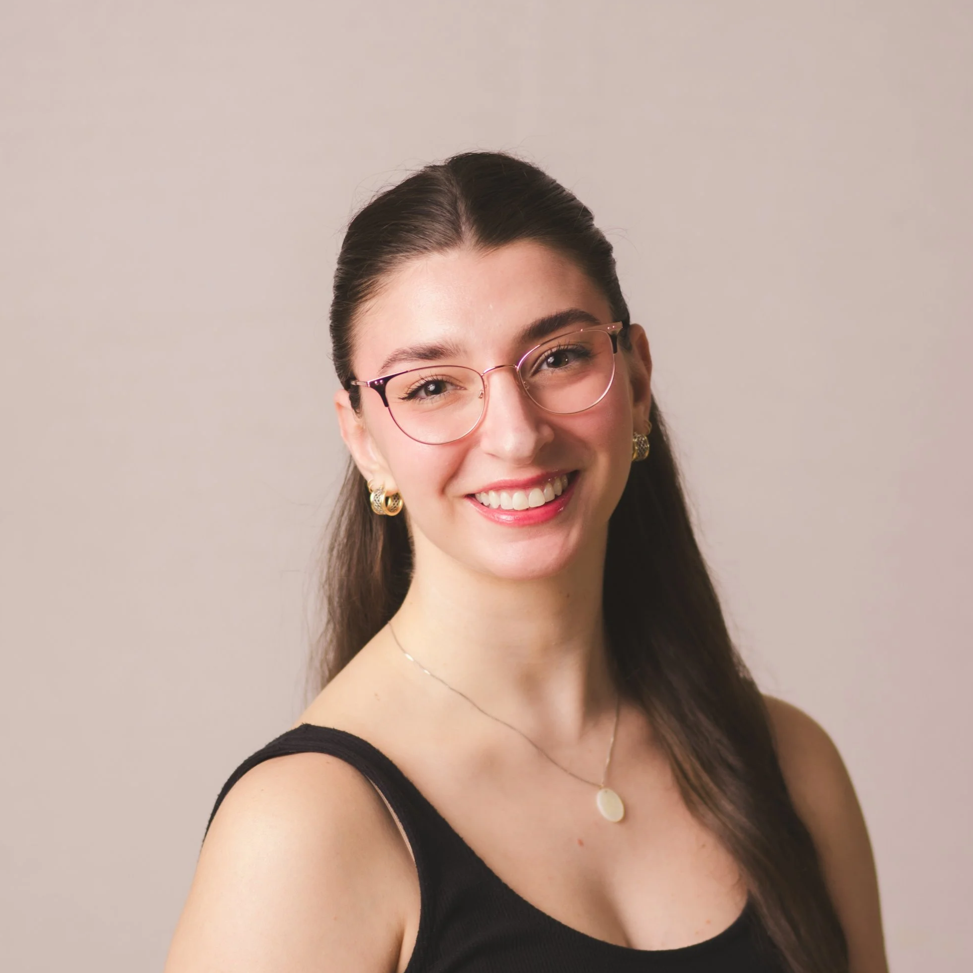 A young woman with shoulder-length brown hair, wearing a black tank top, gold hoop earrings, and a silver necklace, smiling against a plain white background.